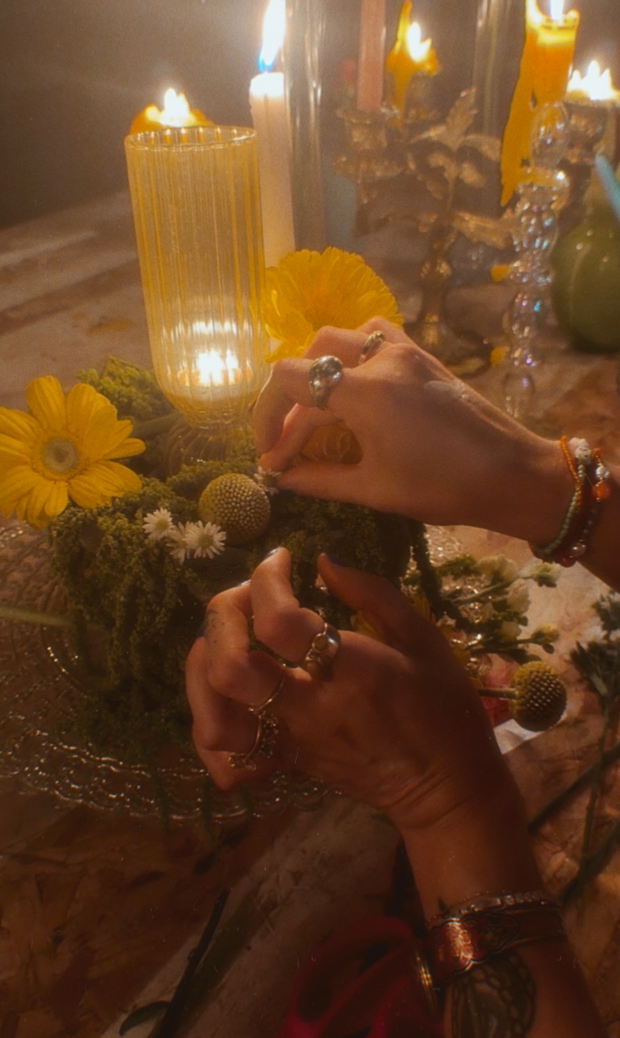 Person's hands with rings and bracelets holding a small object, surrounded by candles, flowers, and decorative items on a table.