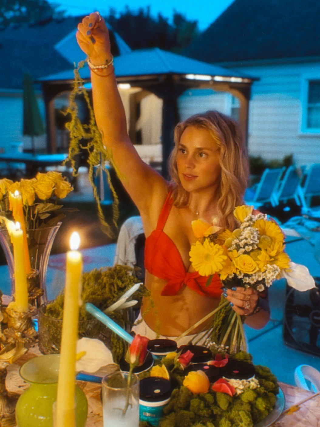 Woman in red top holding a flower bouquet outdoors at evening, surrounded by candles and food, with a house in background.