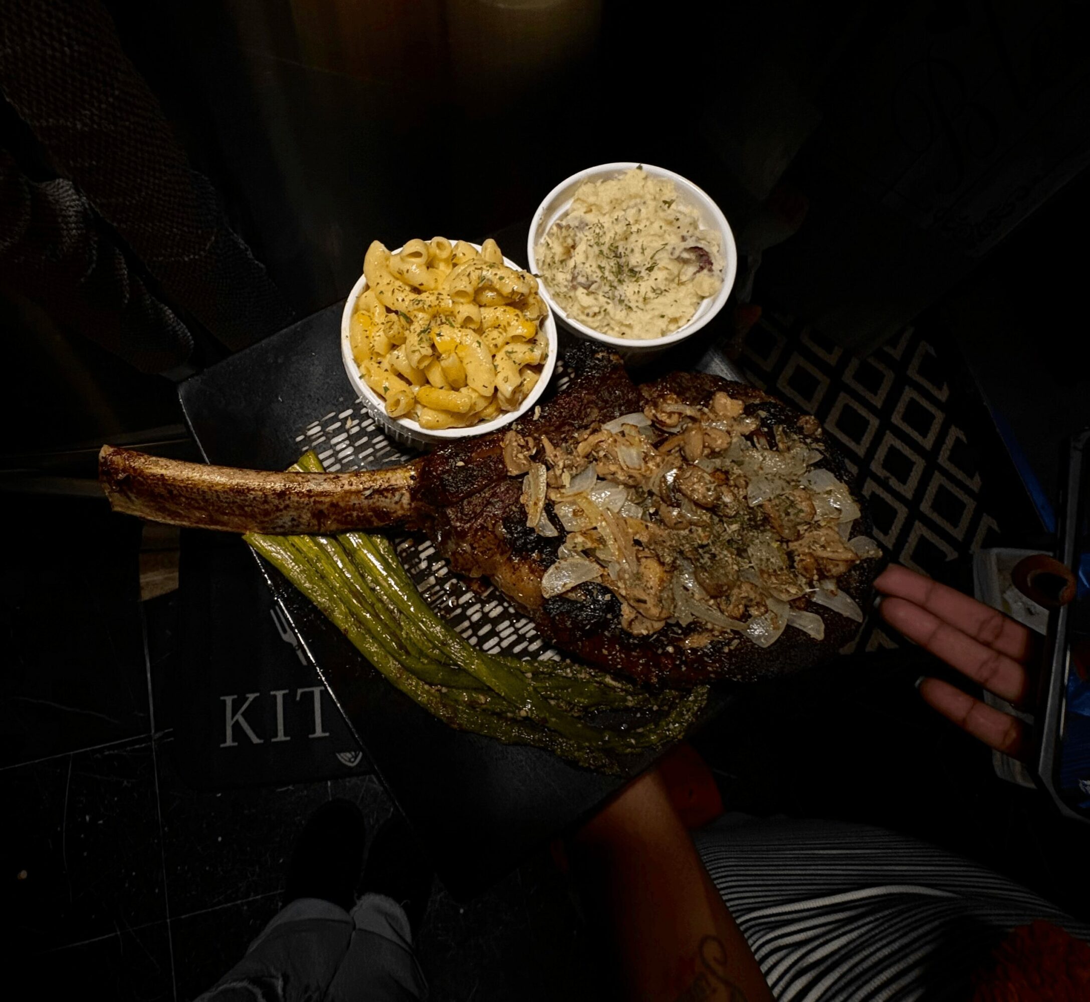Plate with grilled meat, two bowls of side dishes, and a corn cob on a dark surface, viewed from above.