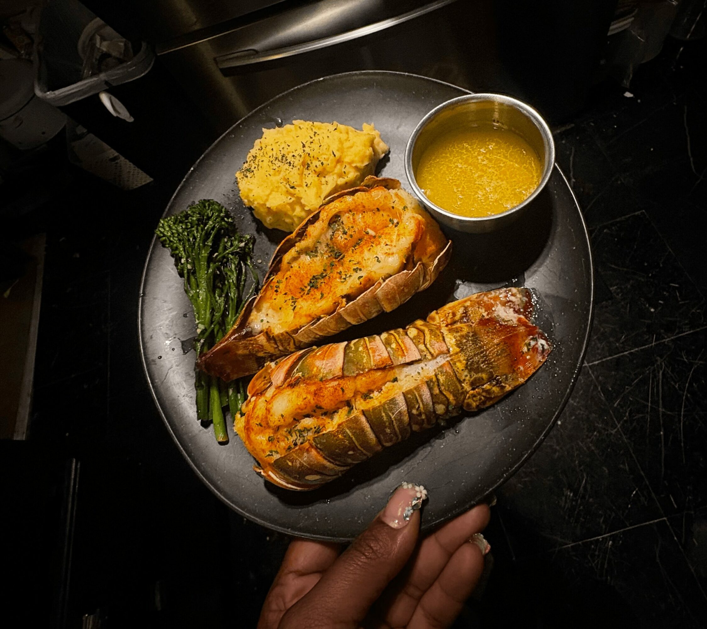 Plate with two lobster tails, broccoli, a piece of bread with topping, yellow sauce in a small cup, and a hand holding the plate.