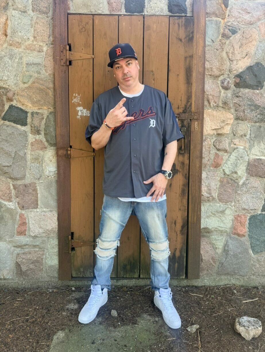 Young man standing in front of a wooden door, wearing a cap, dark shirt, ripped jeans, and white sneakers, making a hand gesture.
