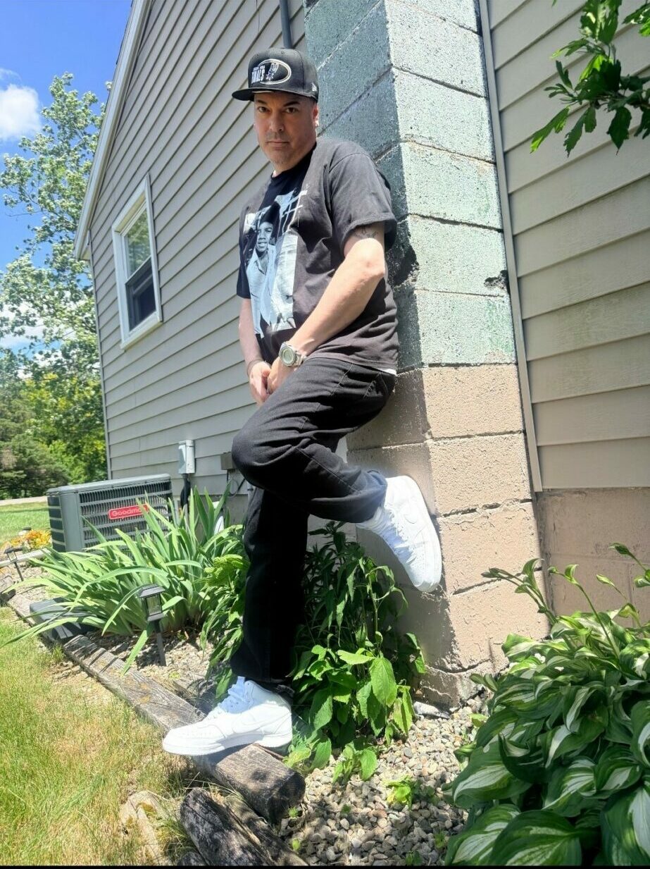 Person leaning against a house wall outdoors, wearing a cap, t-shirt, and pants, with plants nearby.