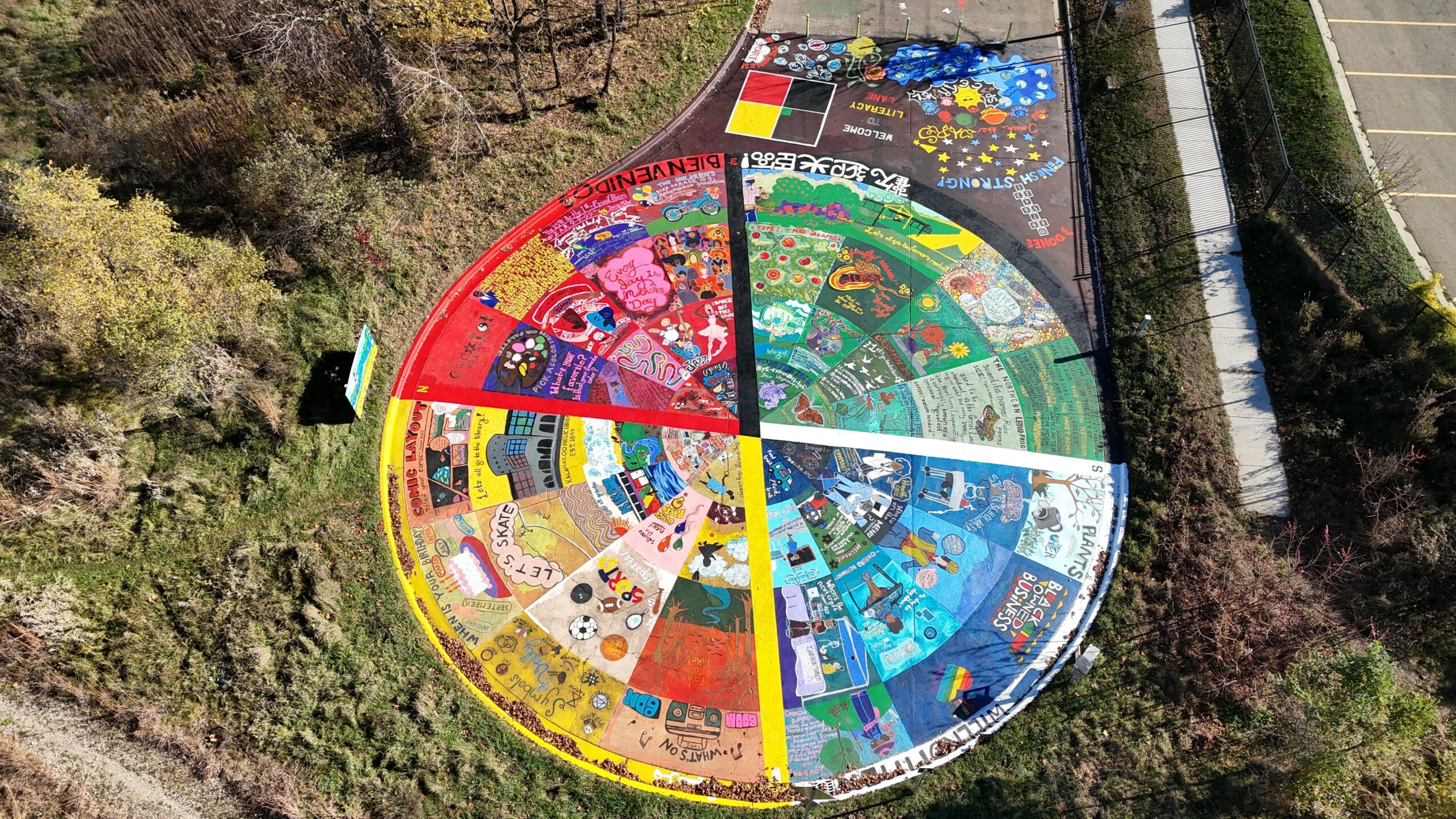 Colorful circular mural divided into sections, painted on the ground outdoors, surrounded by trees and a fence.