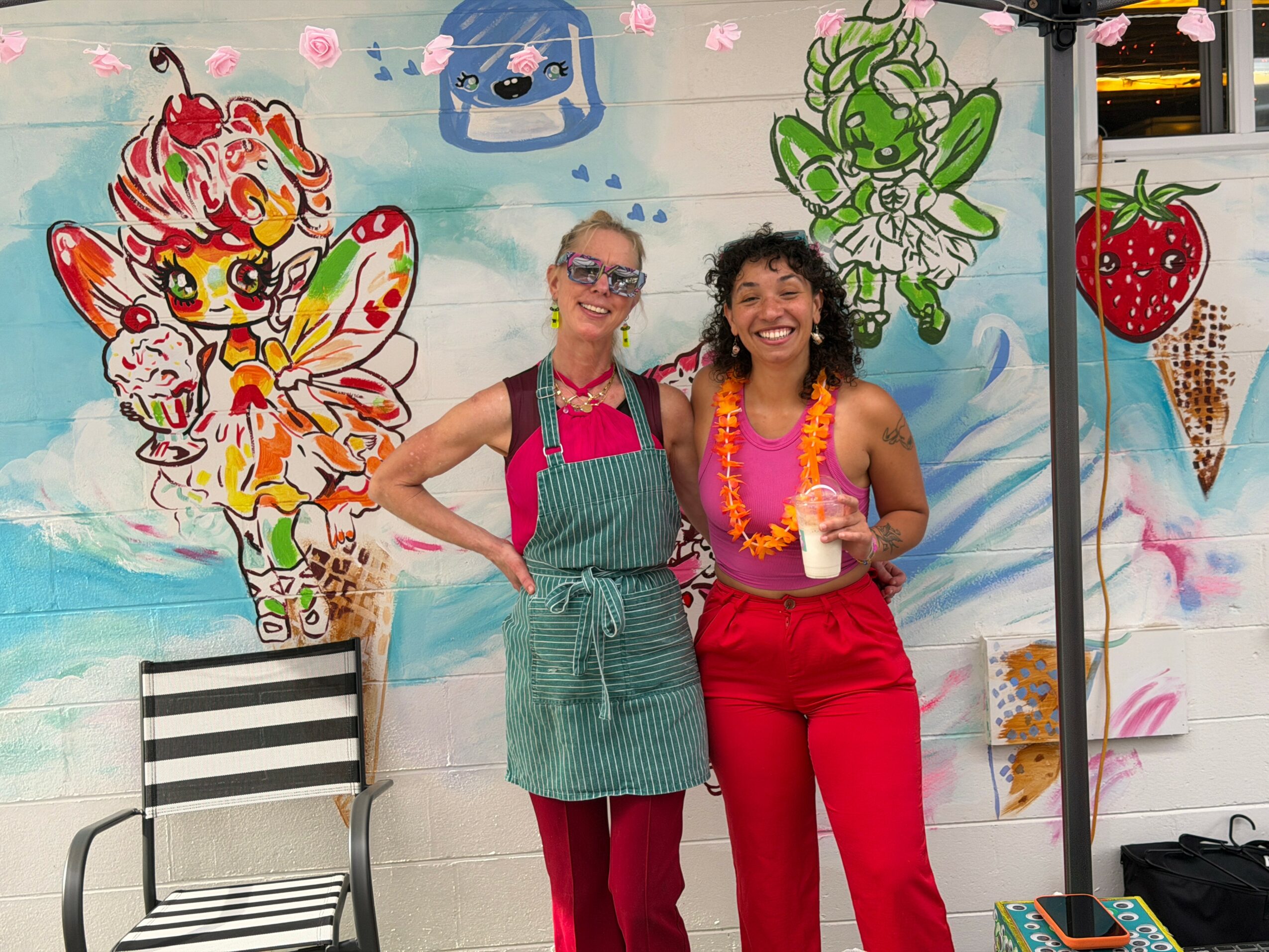 Two women smiling in front of a colorful mural, one holding a drink, both wearing aprons and bright clothing.