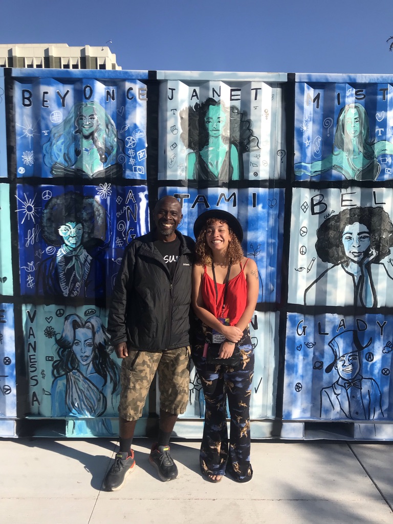 Two people stand in front of a mural with portraits and text, smiling outdoors on a sunny day.