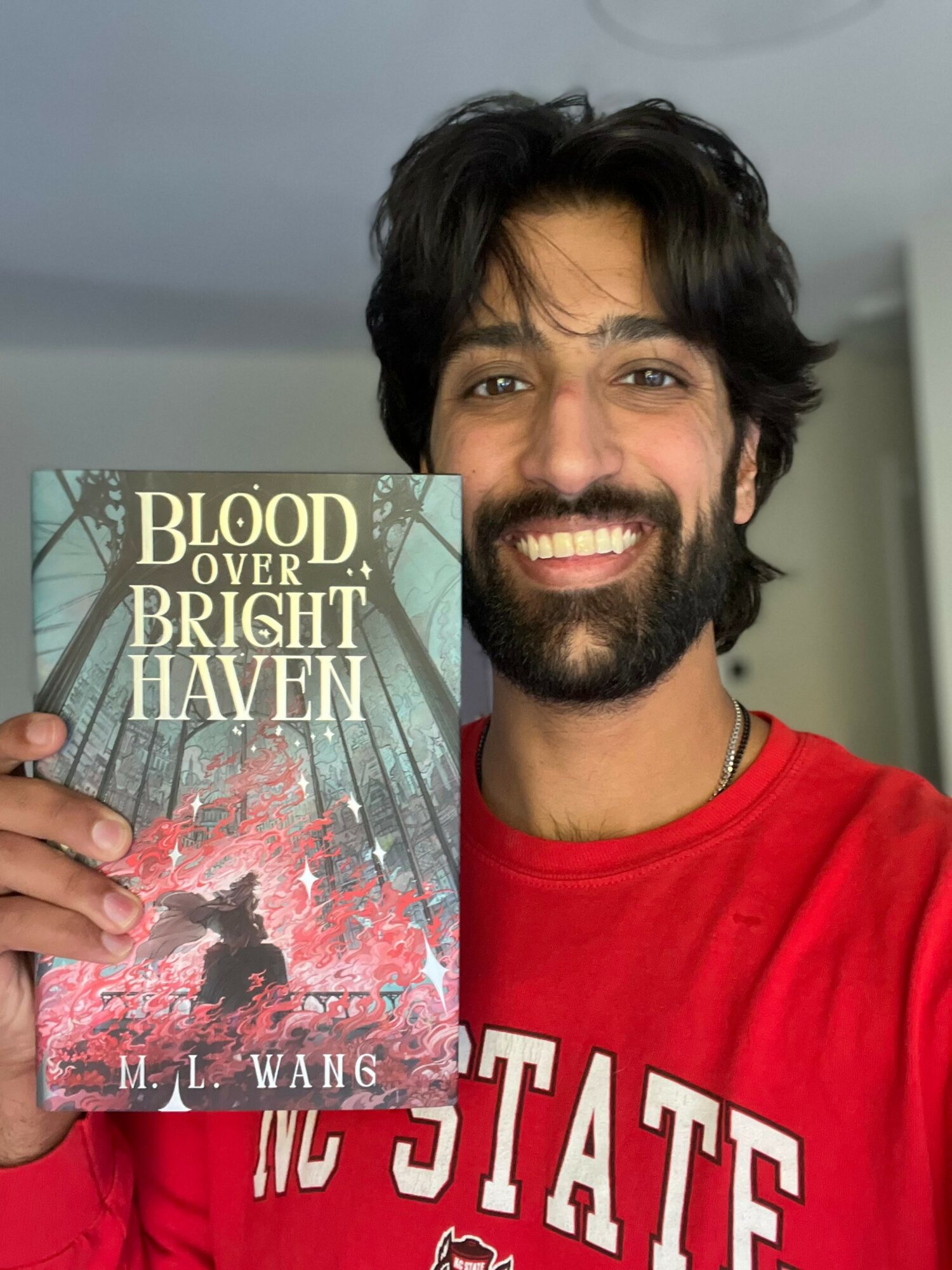 Man with dark hair and beard smiling, holding a book titled 'Blood Over Bright Haven' by M. L. Wang.