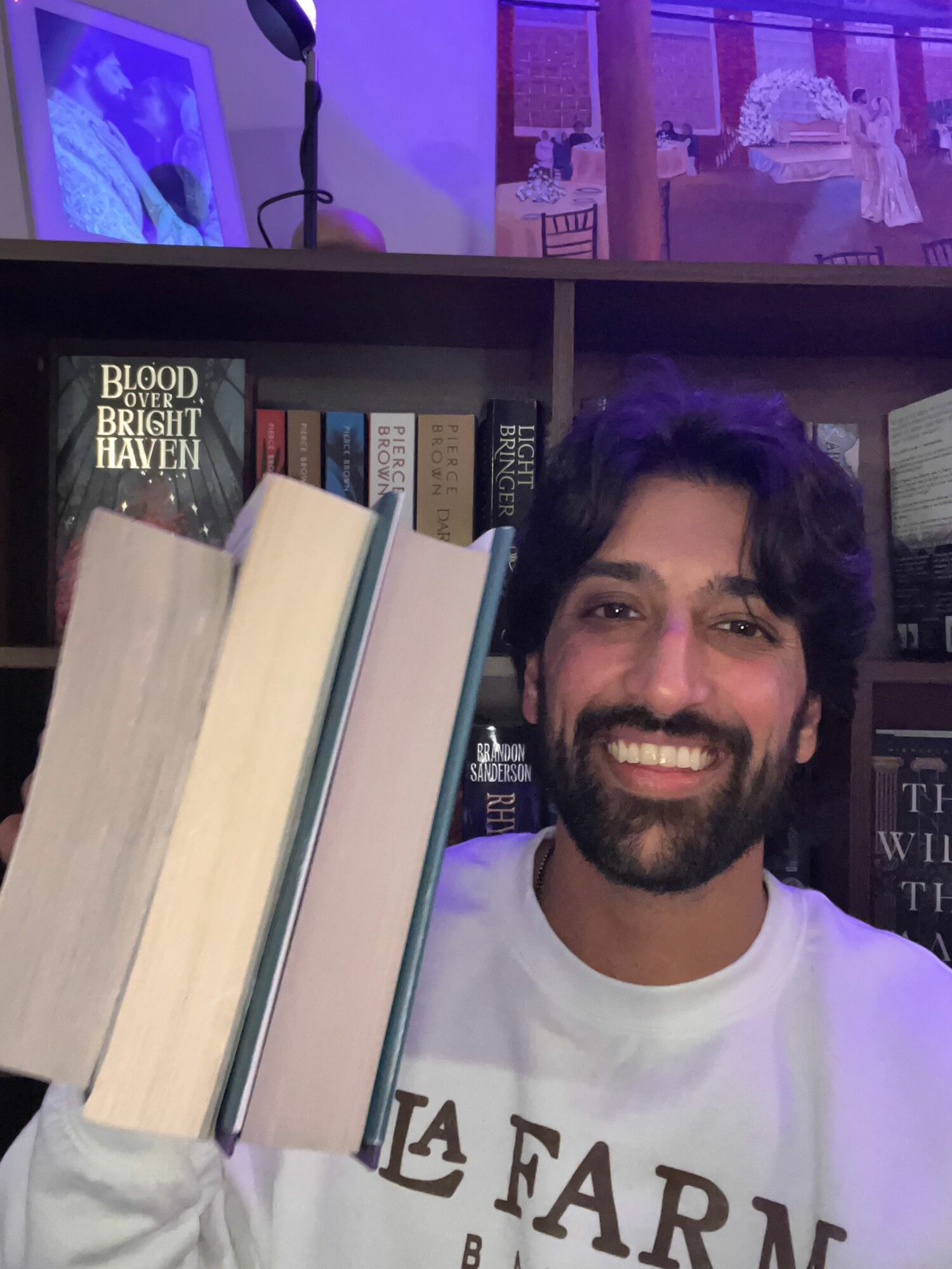 Smiling man with dark hair and beard in front of bookshelf, holding two large books, wearing a white shirt.