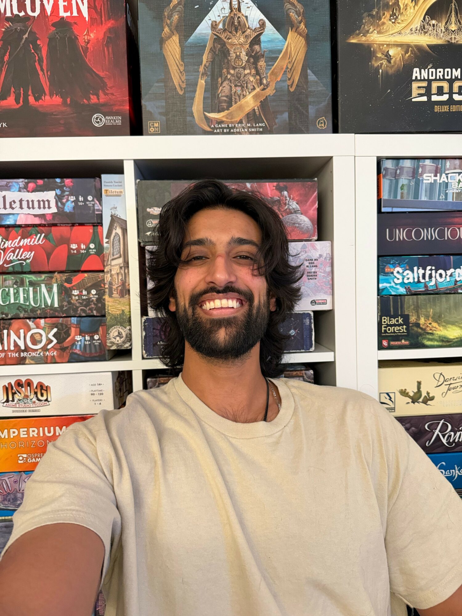 Man with long dark hair and beard smiling, wearing a light-colored shirt, in front of a bookshelf filled with books.