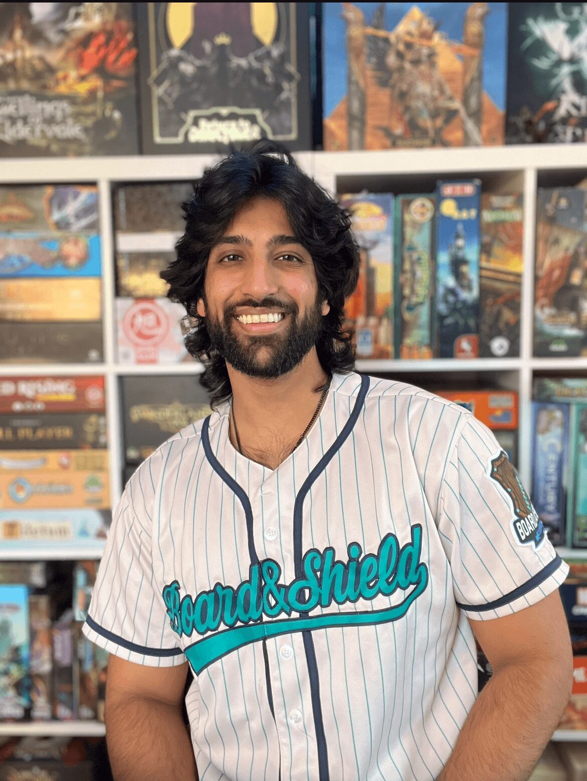Smiling man with dark curly hair and beard wearing a baseball jersey in front of a game shelf.