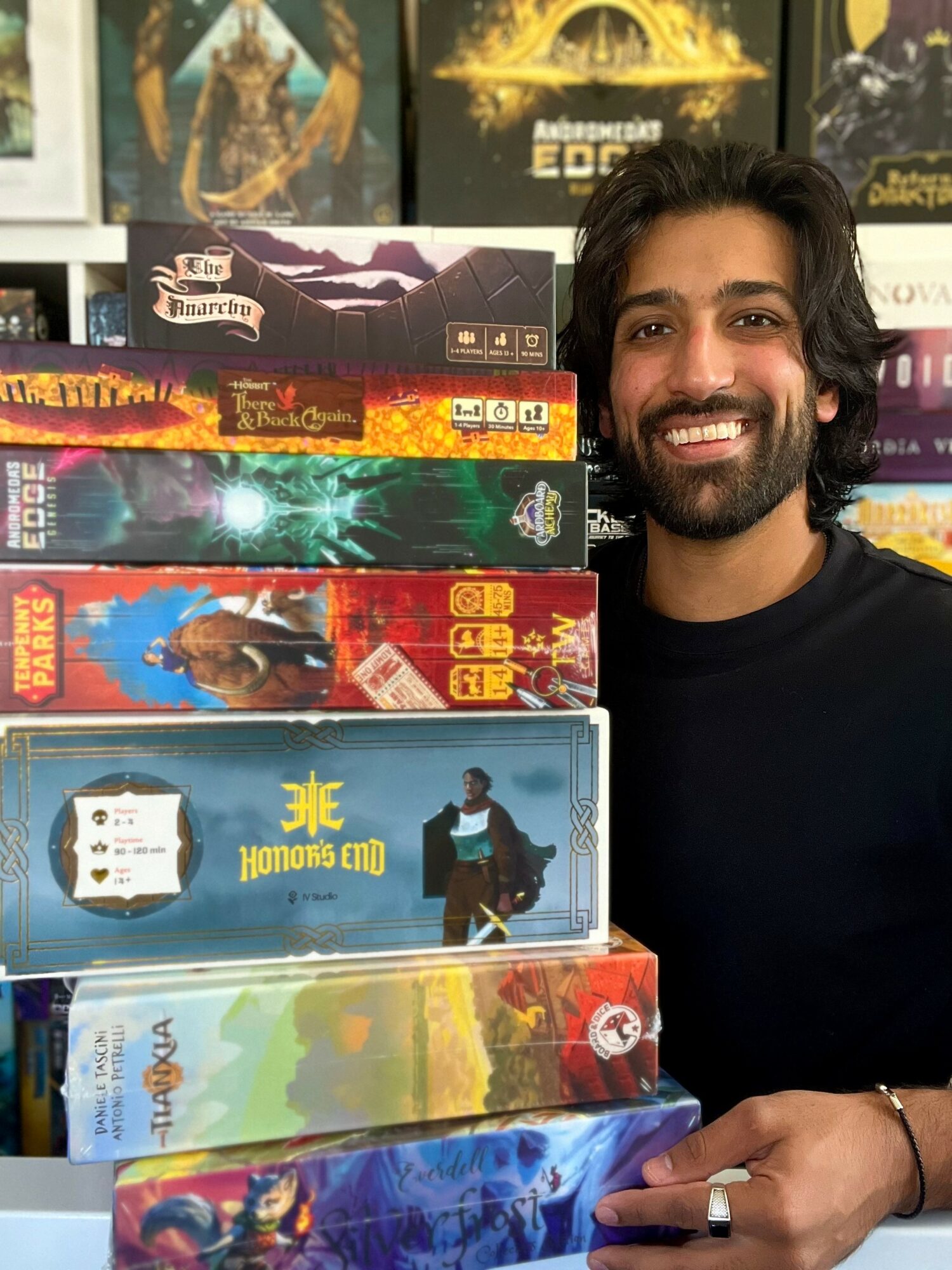 Man with dark hair and beard smiling next to a stack of colorful board games and books.