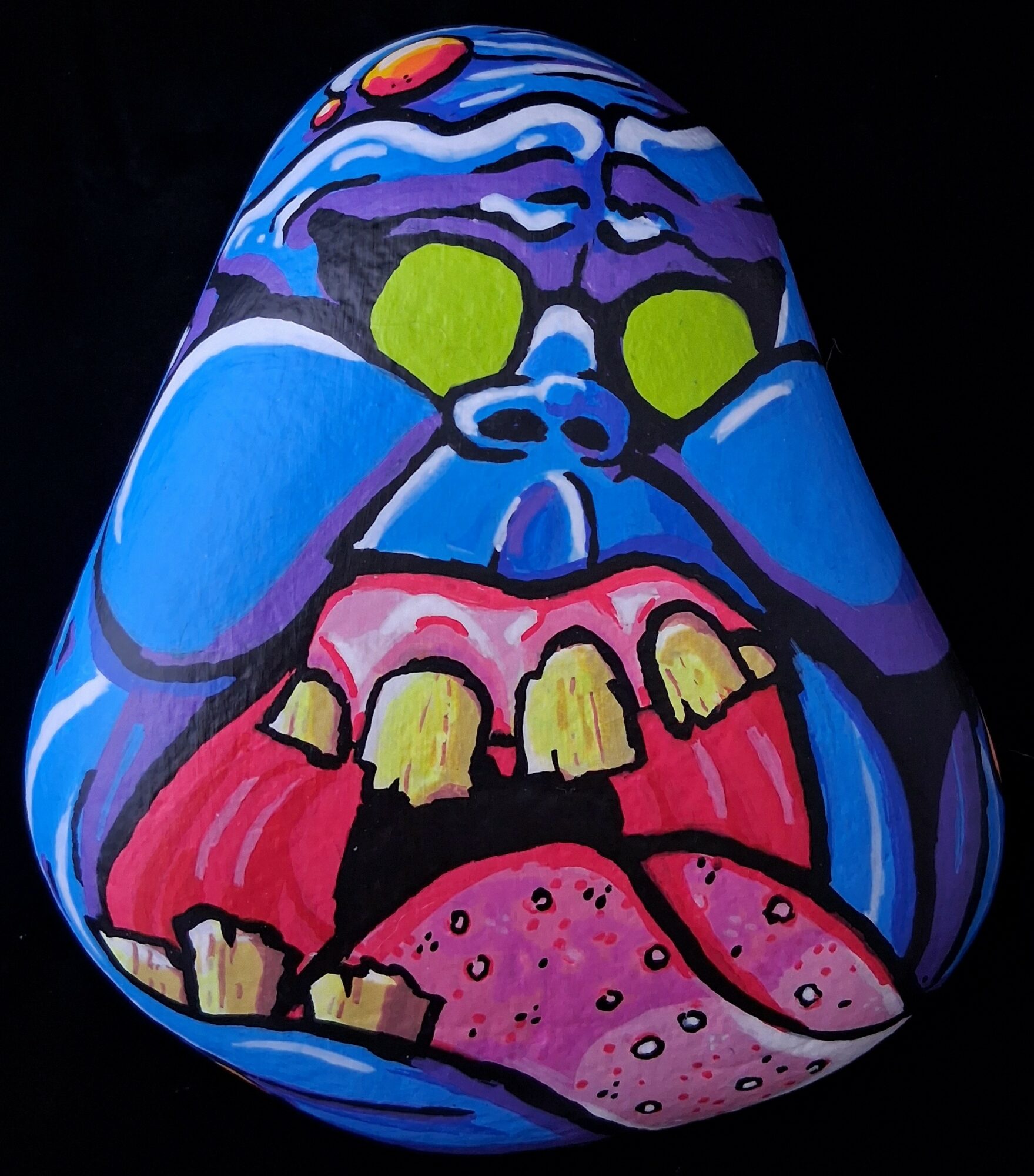 Colorful painted rock with a face, tongue sticking out, and sunglasses, on a black background.