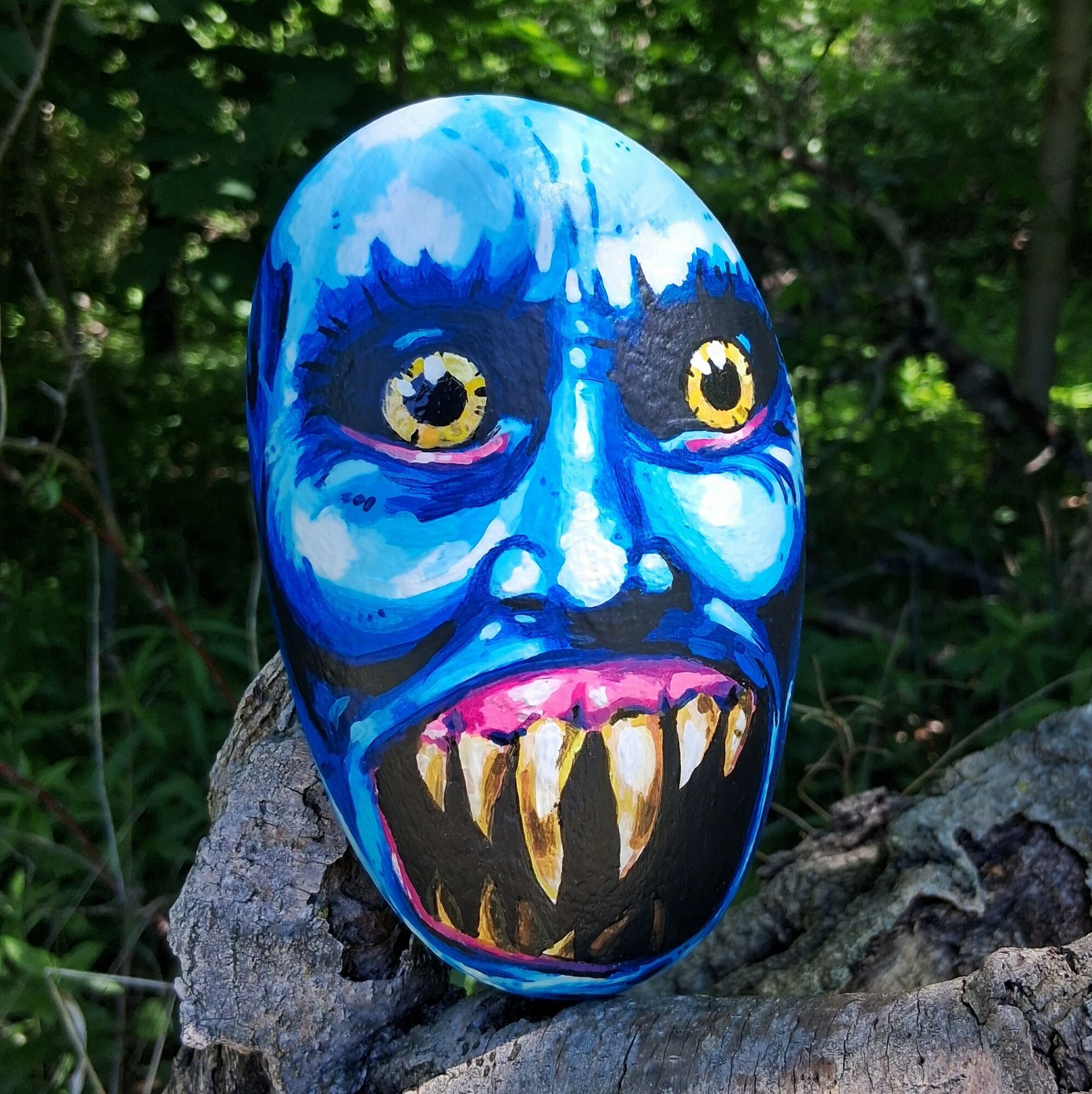 Colorful painted mask with blue face, yellow eyes, and sharp teeth, resting on a tree trunk outdoors.