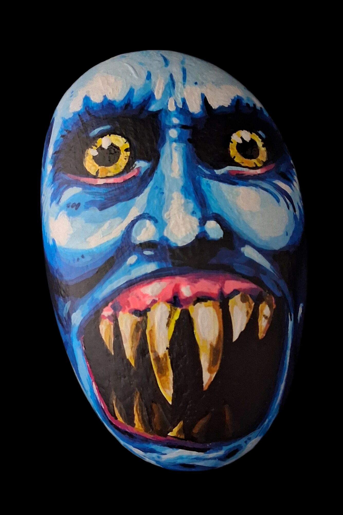Colorful painted mask with exaggerated facial features, sharp teeth, and large yellow eyes, set against a black background.