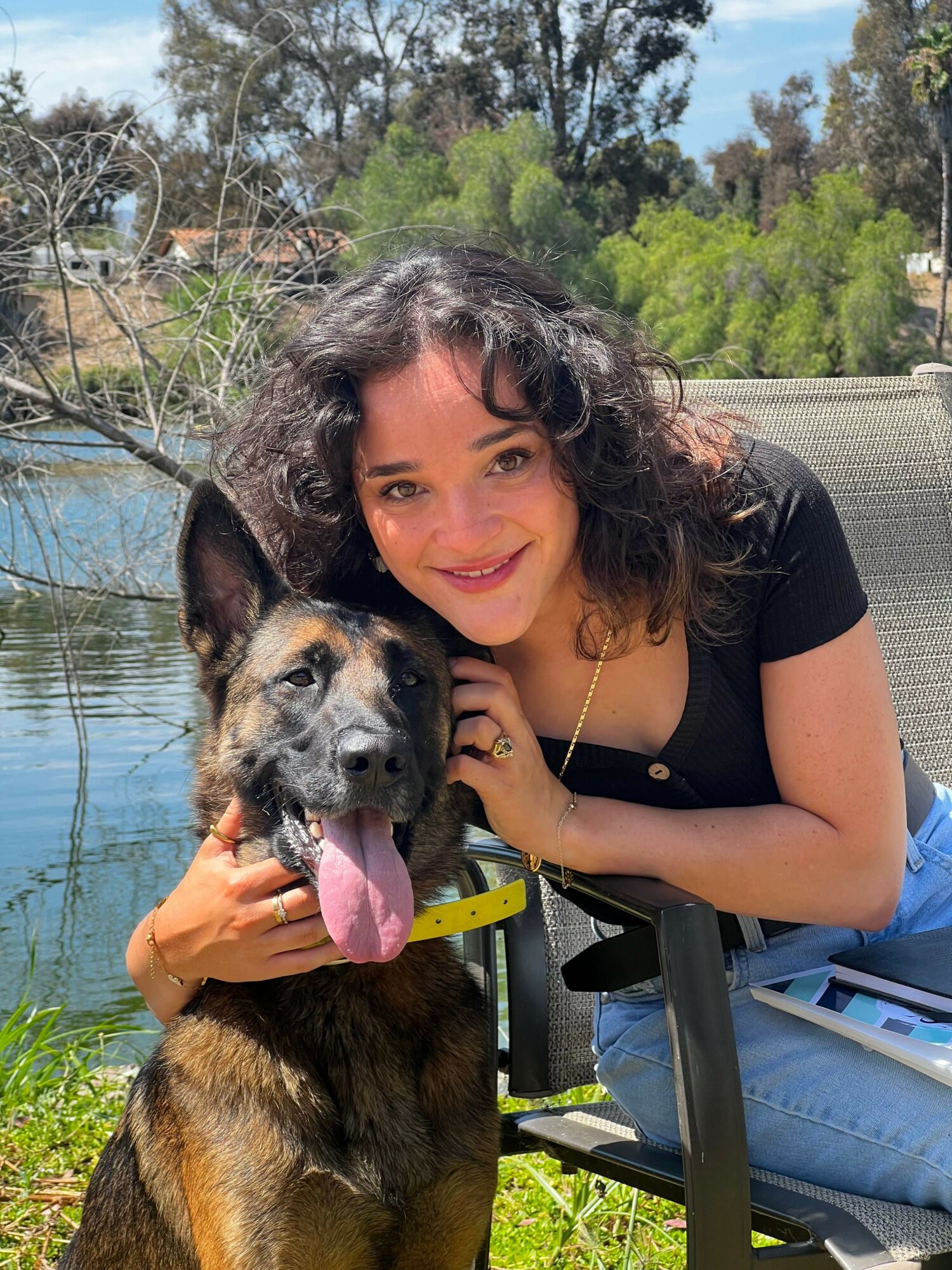 Woman with curly hair and black shirt holding a large dog outdoors near water, trees in background.