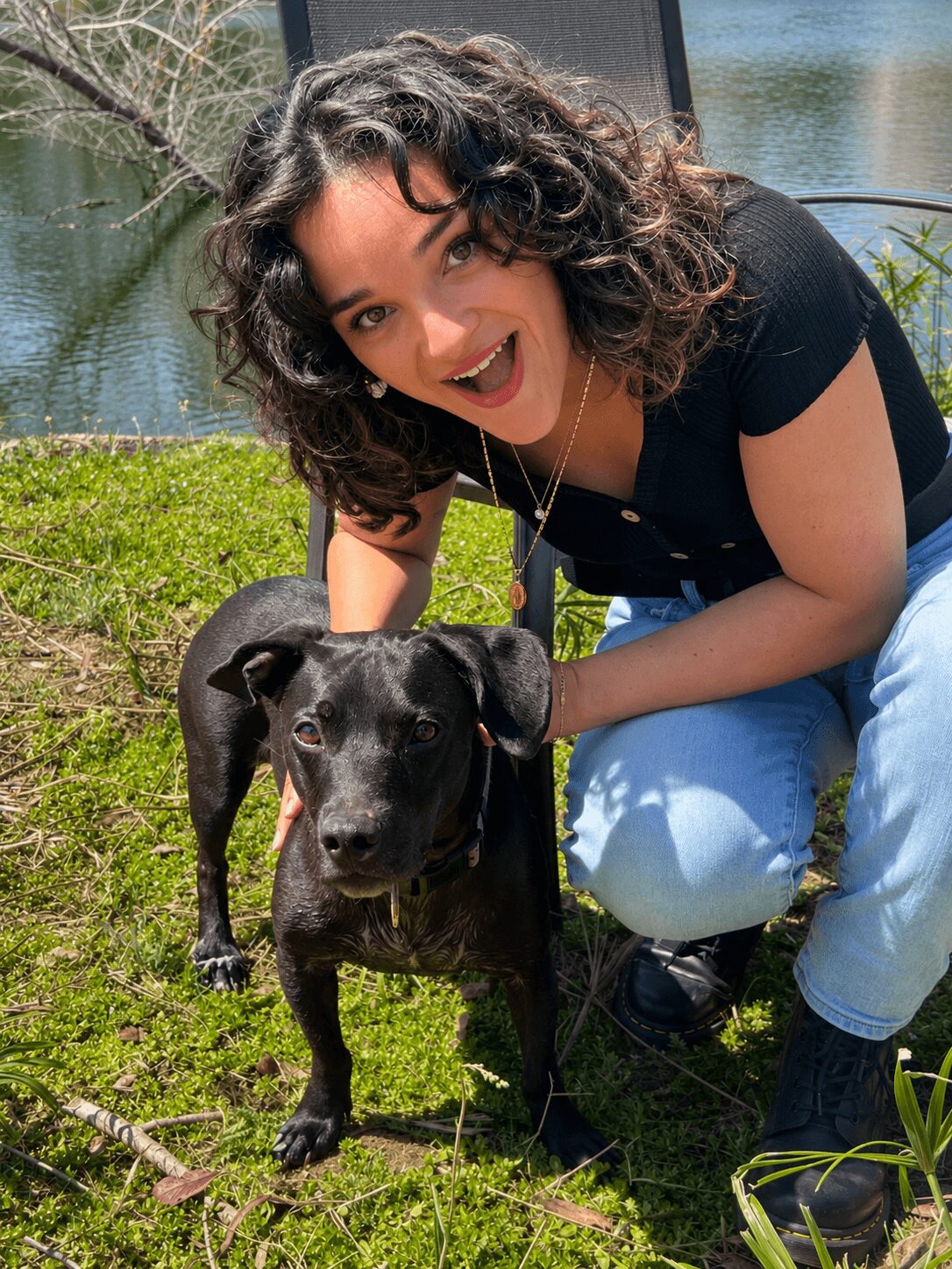 Young woman with curly hair smiling outdoors next to a black dog on grass near water.