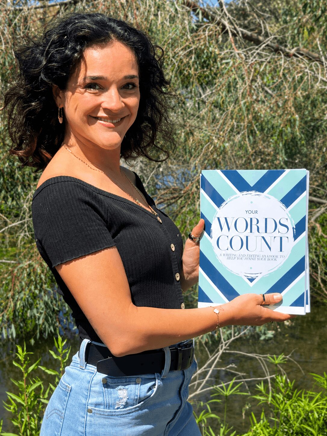 Woman with curly dark hair holding a book outdoors, smiling, with trees and greenery in background.