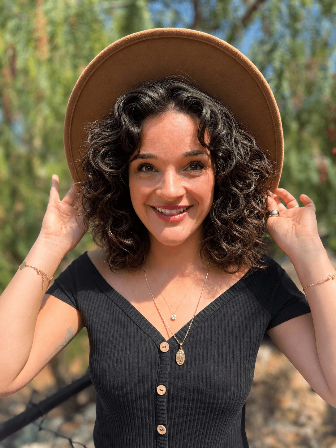 Young woman with curly hair wearing a wide-brimmed hat outdoors, smiling, holding the hat with both hands, trees in background.