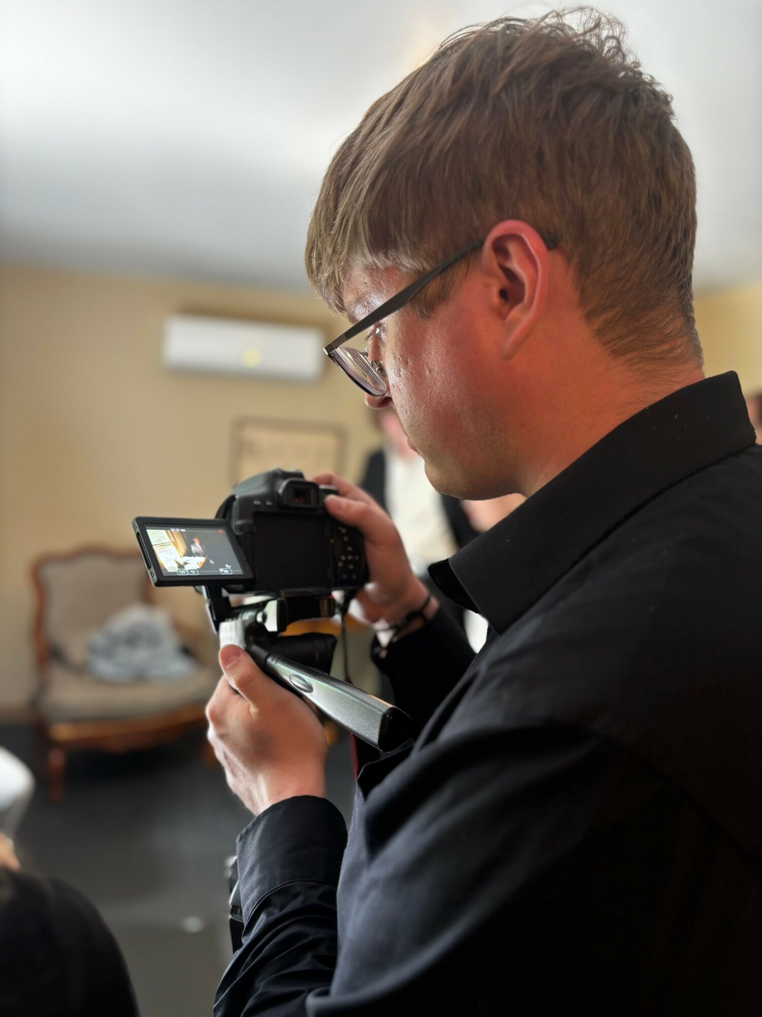 Man with glasses and short hair looking at a camera, holding a smartphone, in an indoor setting.