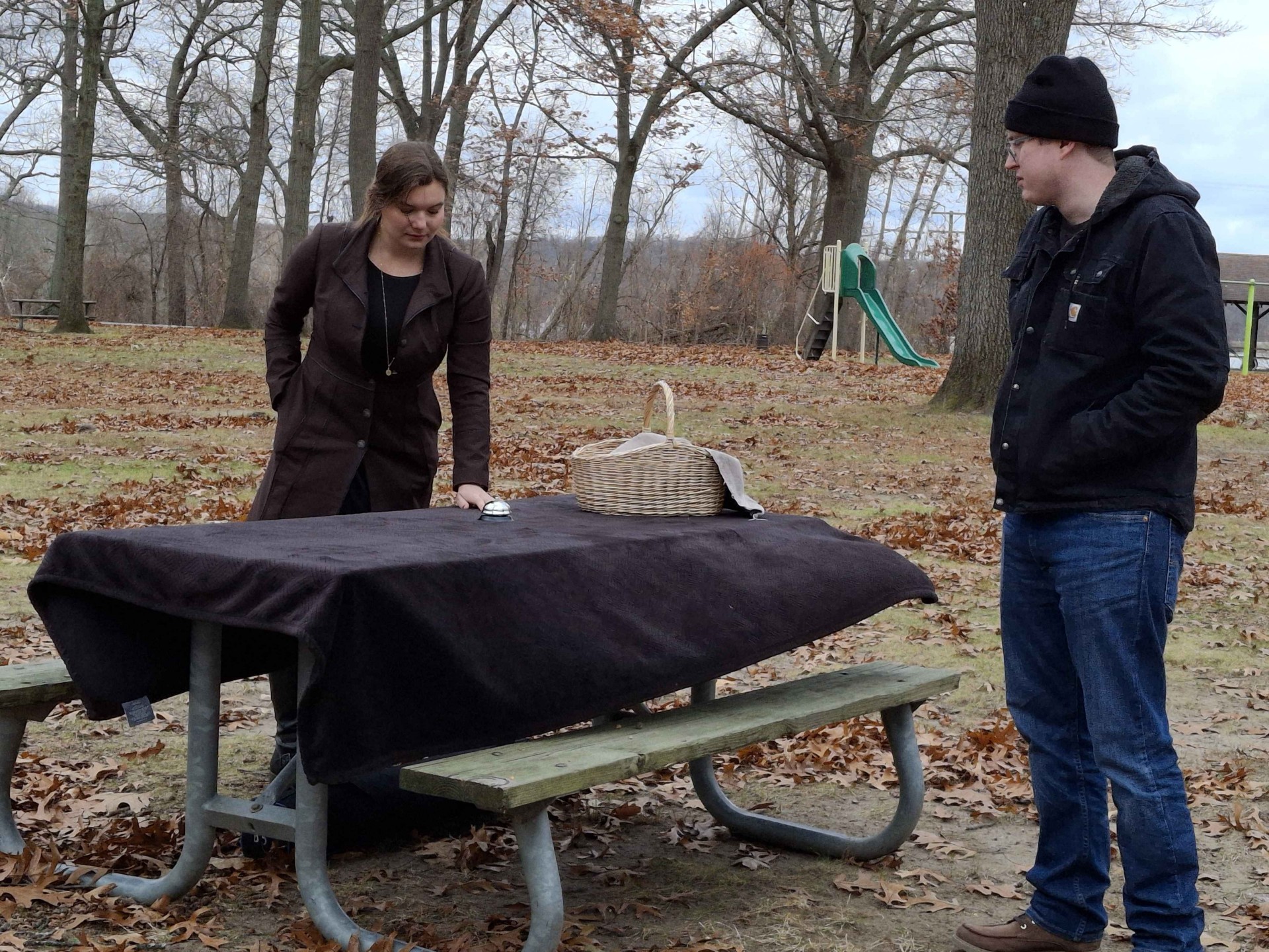 Two people stand near a picnic table outdoors in a park with trees and a slide in the background.