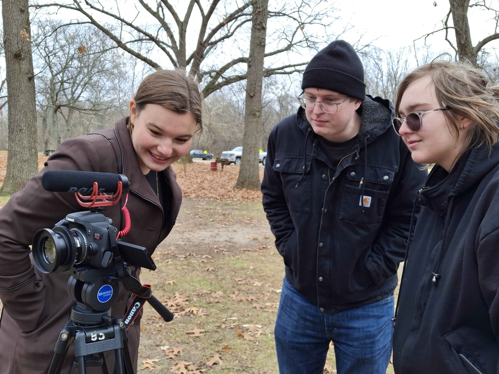 Three people outdoors, two women and one man, looking at a camera on a tripod, with trees and fallen leaves in the background.
