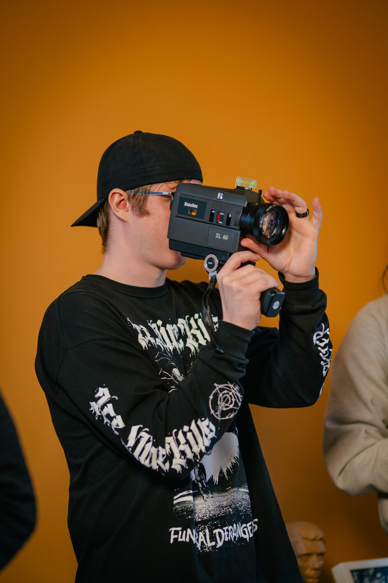 Person wearing a black cap and black sweatshirt holding a vintage camera, against an orange background.