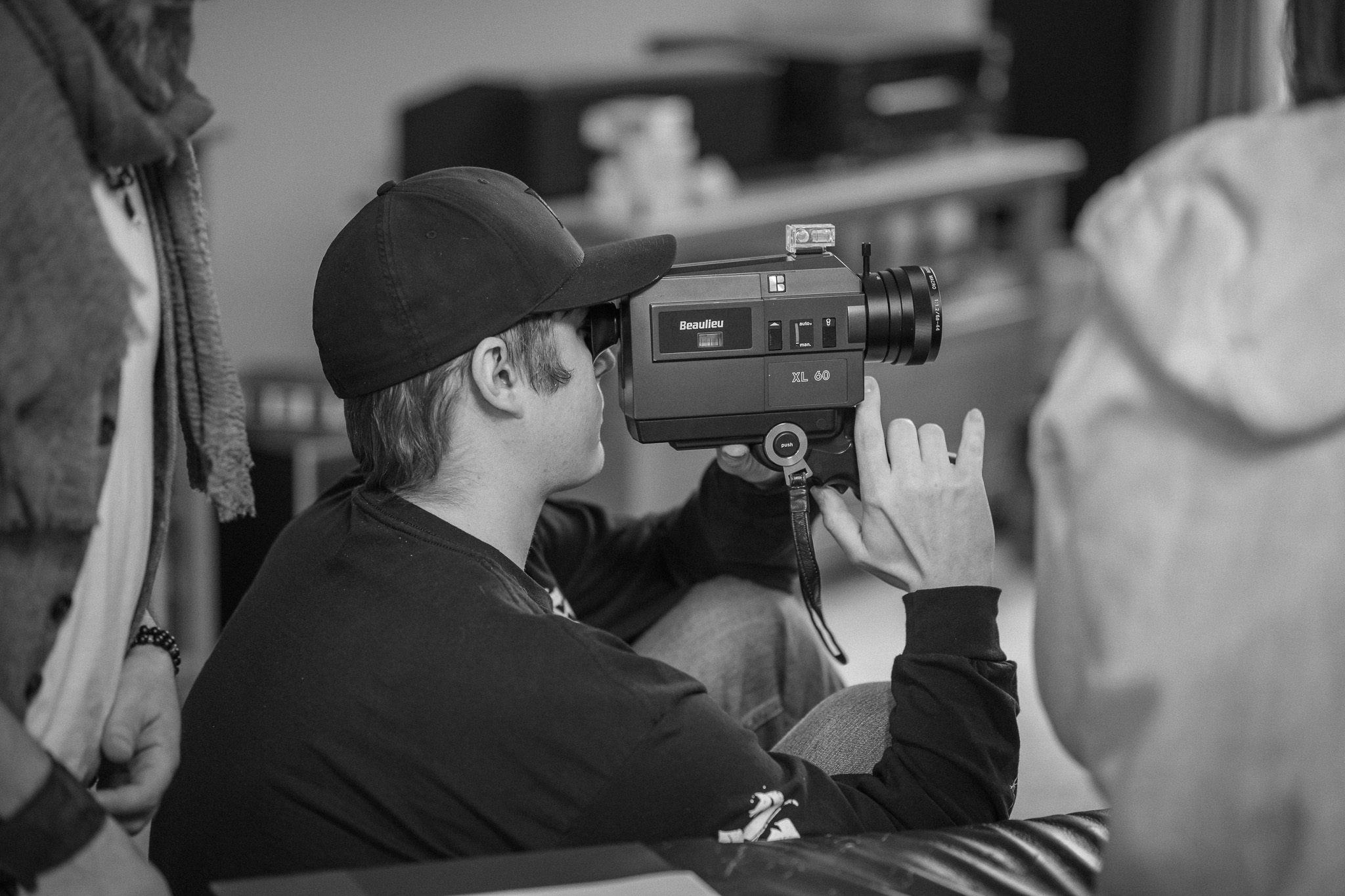 Child wearing a cap uses a video camera indoors, with people and furniture in the background.