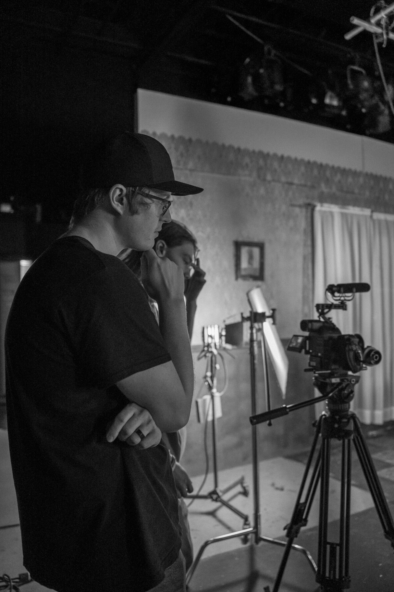 Person wearing glasses and a cap, standing near a camera on a tripod, in a dimly lit room with a brick wall.