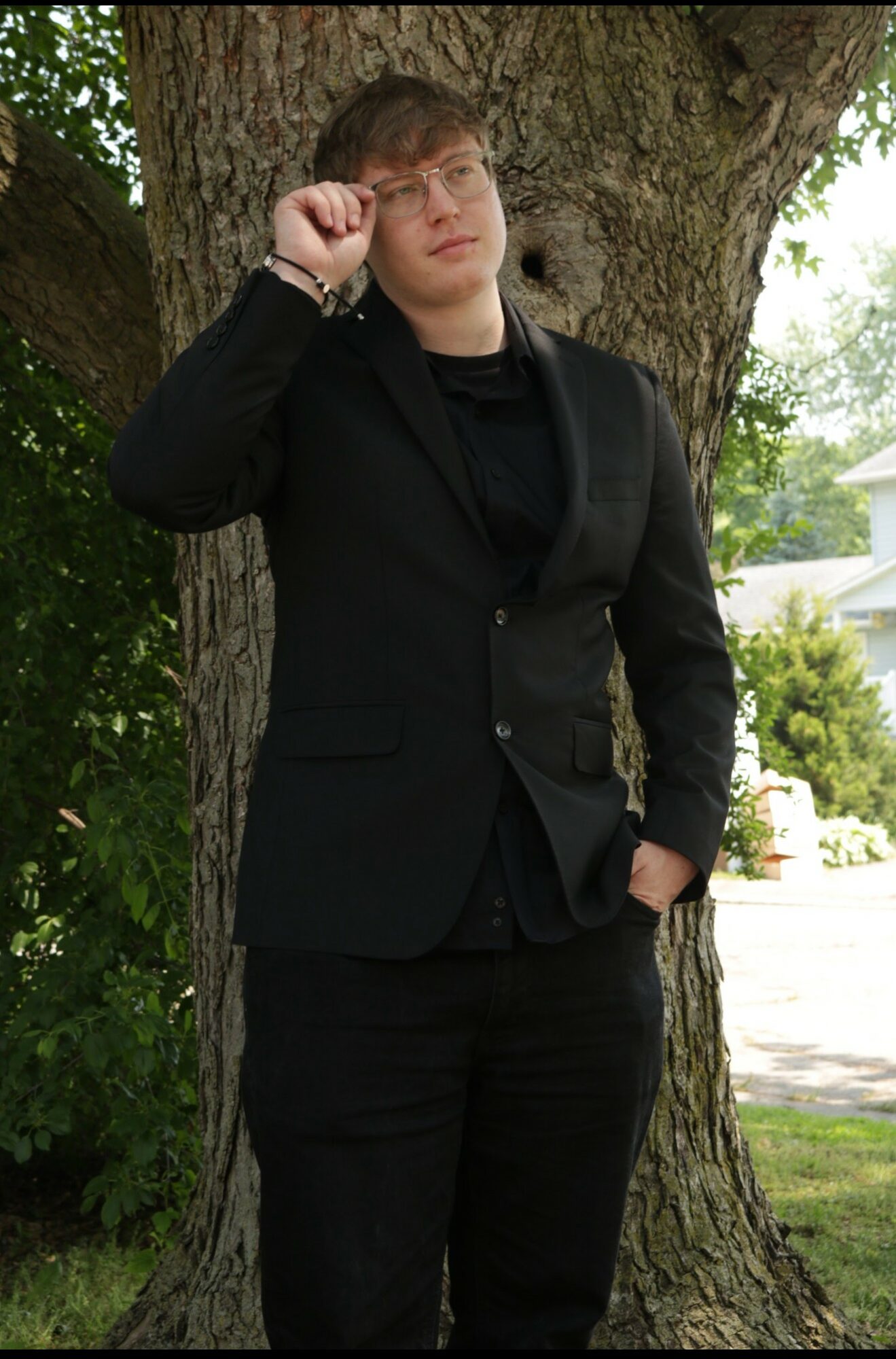 Person in black suit standing outdoors near a large tree, with one hand touching their glasses.