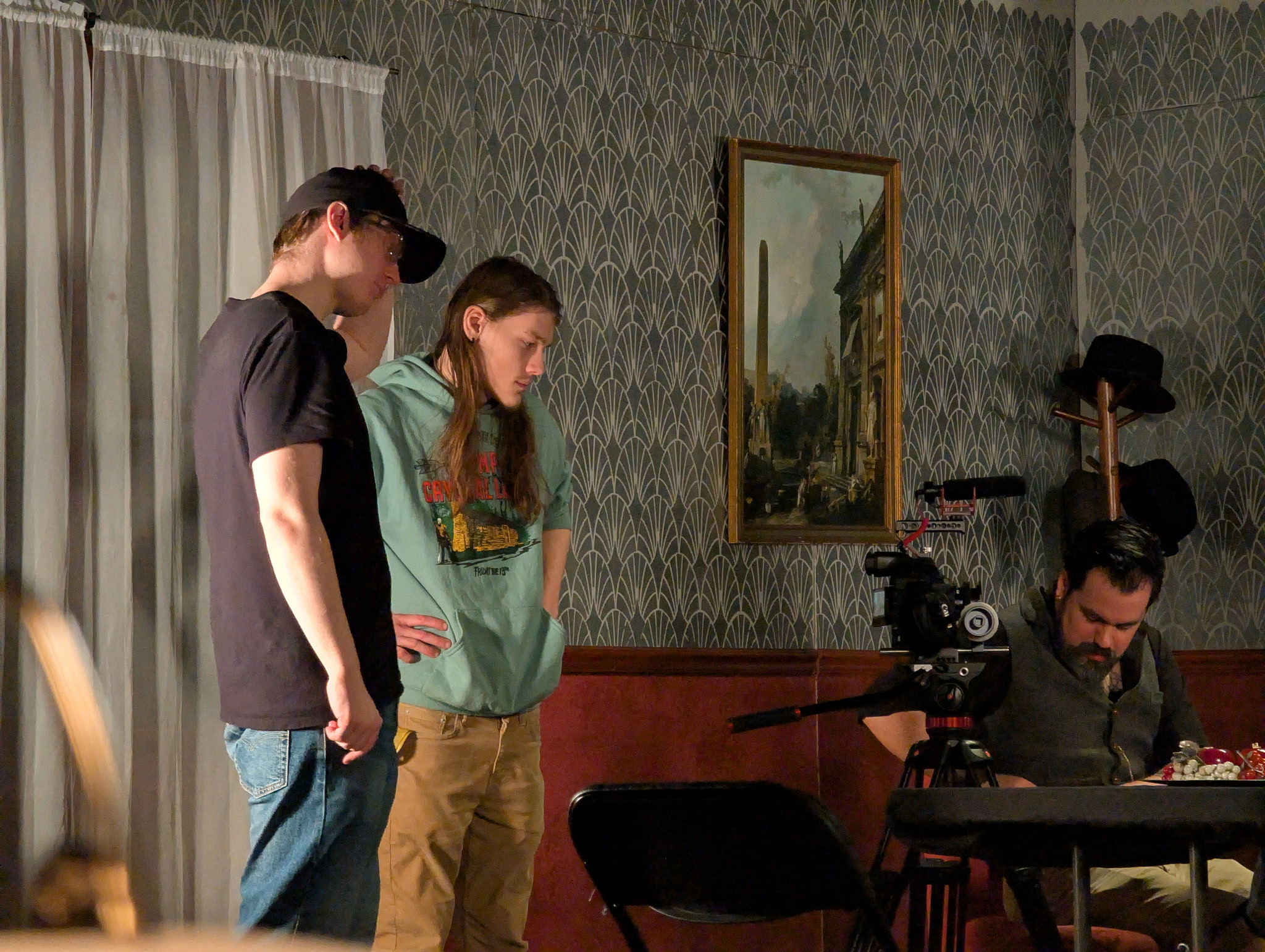 Two people stand watching a man operating a camera at a table in a room with patterned wallpaper and a framed painting.