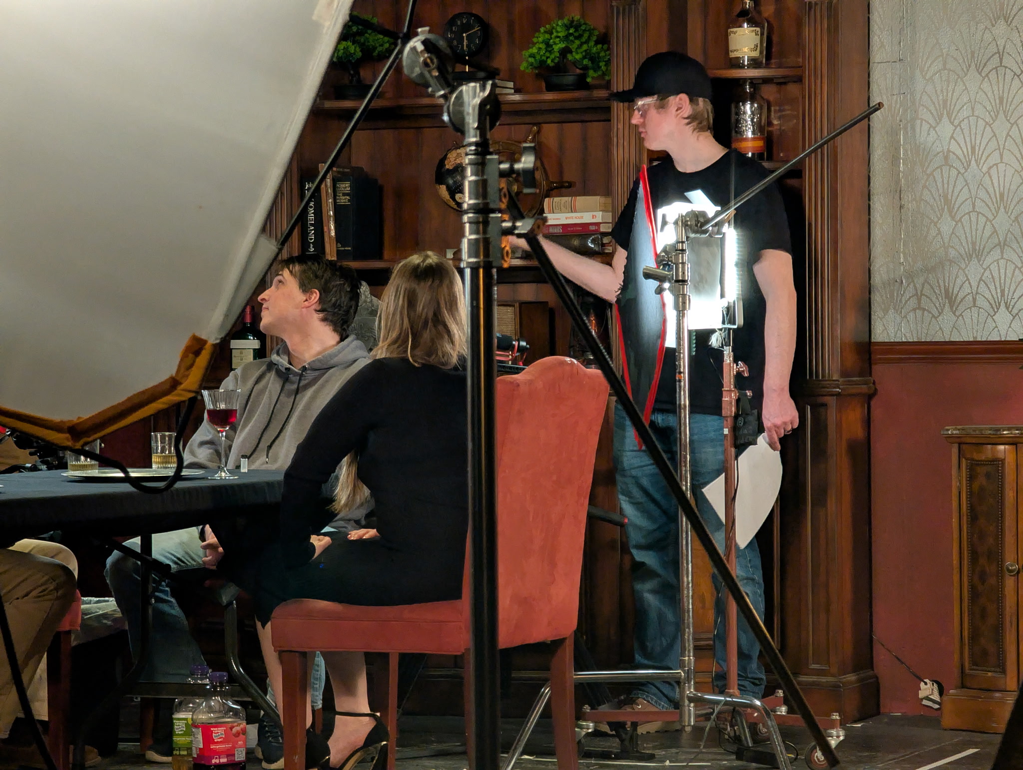 People sitting at a table with a person standing and holding a script, in a room with wooden shelves and decor.