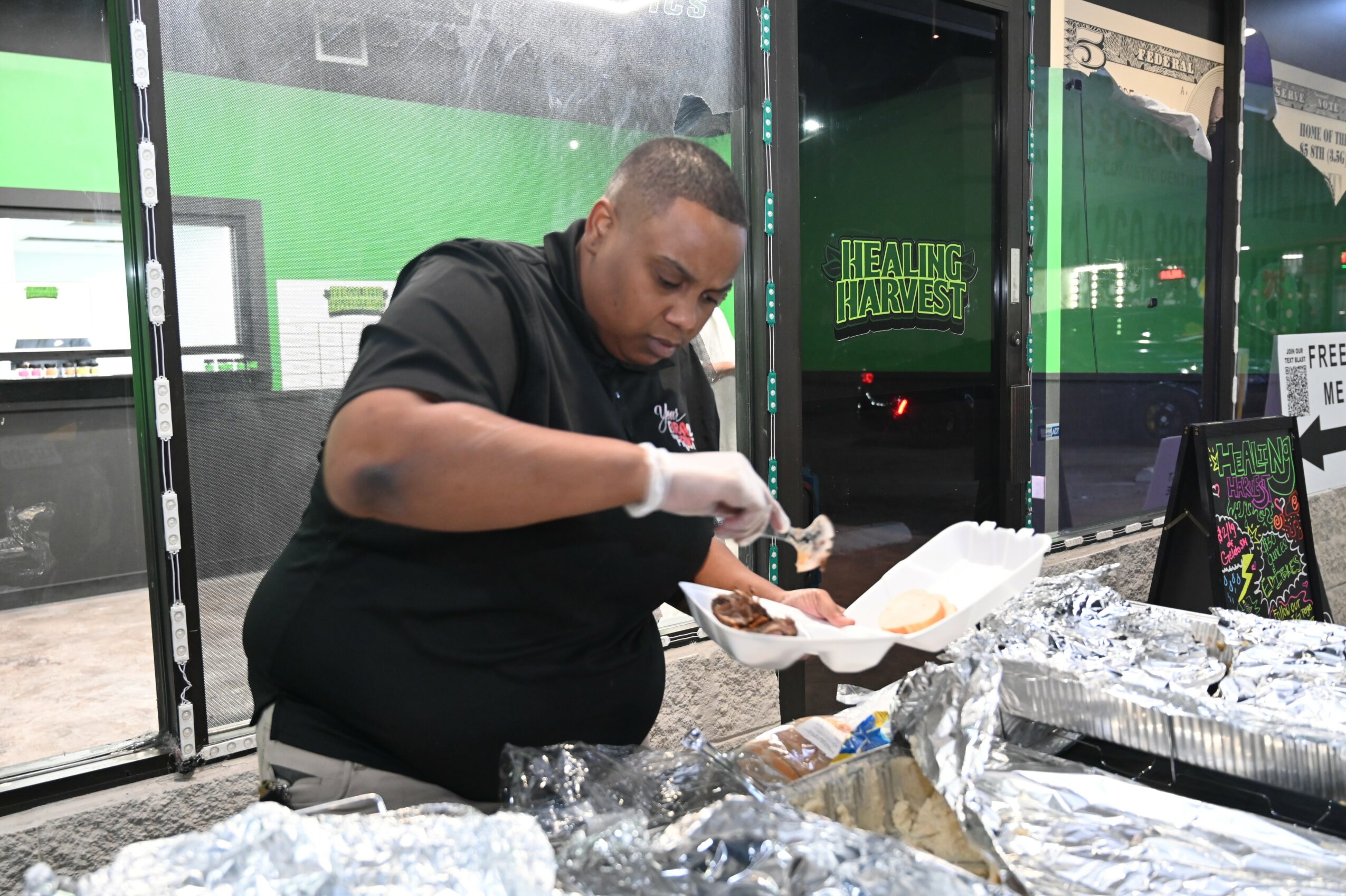 Person serving food at a counter with foil-covered trays, in a setting with green and black decor.