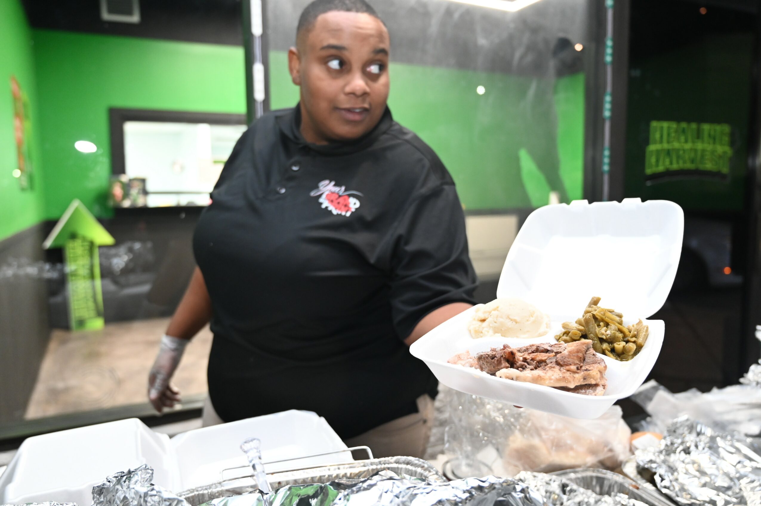 Person holding a takeout container with food, standing behind a counter, in a green and black interior setting.