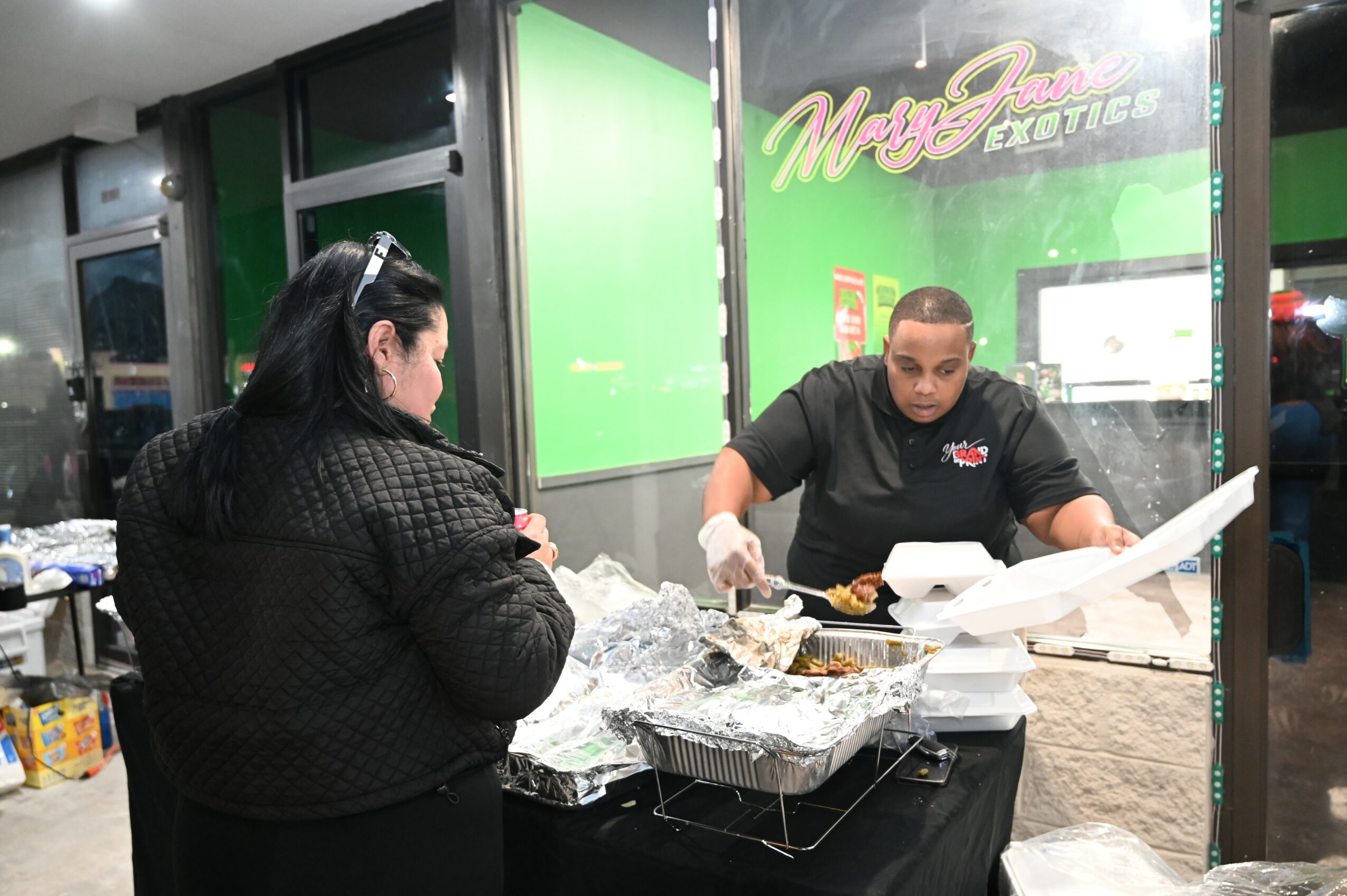 A man serving food at a food stand with a woman customer in front, in an indoor setting.