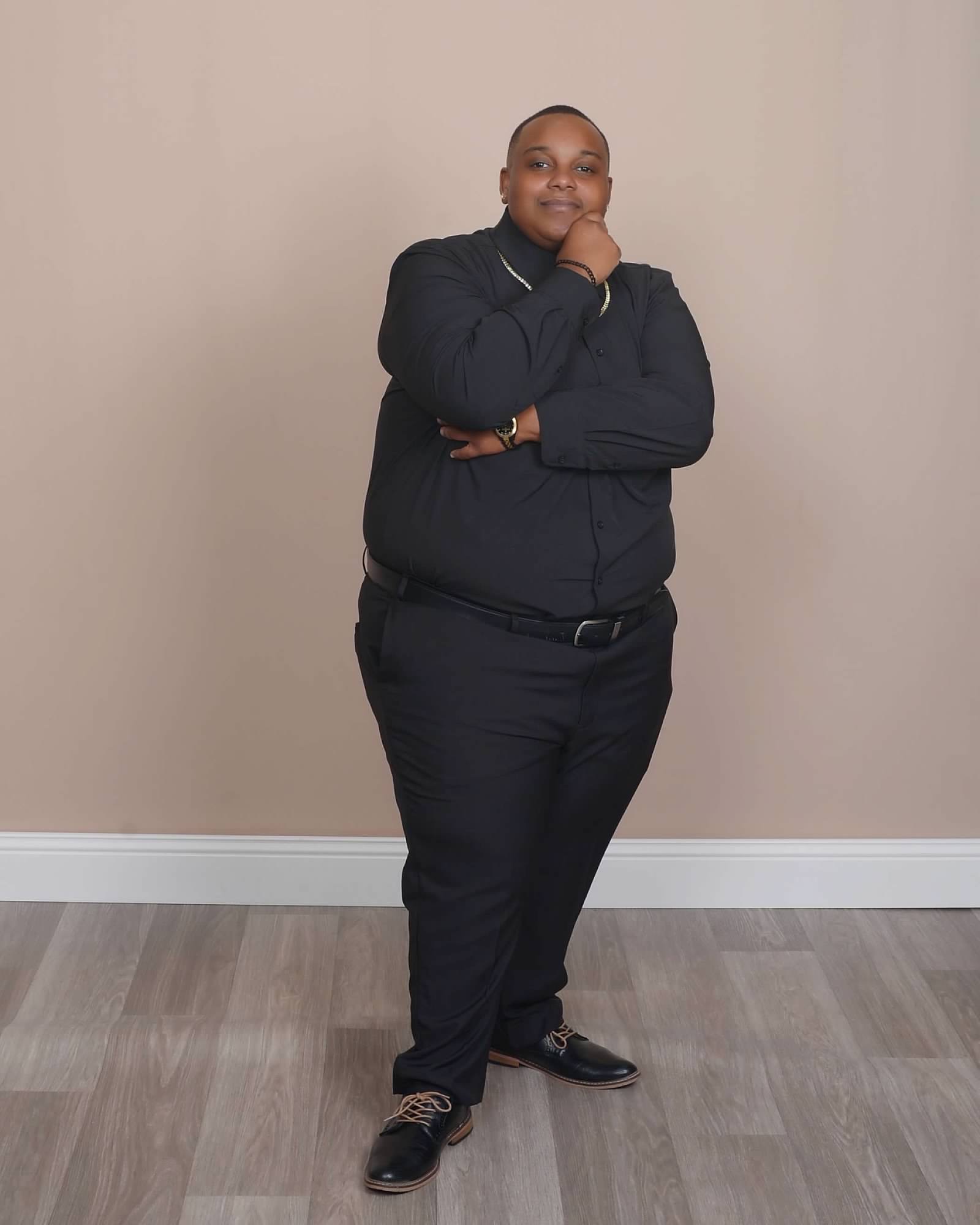 Person standing with hand on chin, smiling, wearing dark clothing, against a plain beige wall.
