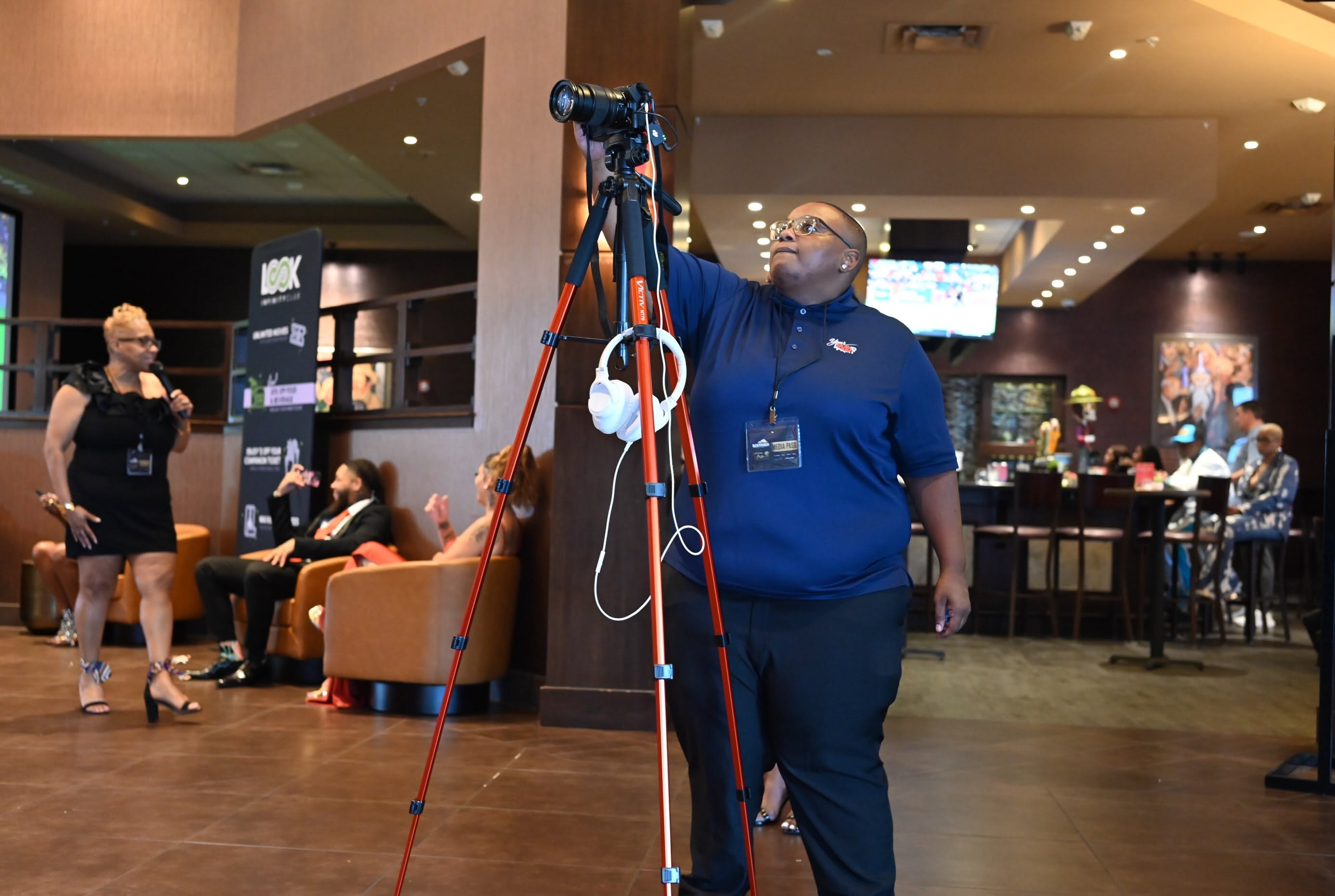 Person adjusting camera on tripod in indoor event space with people and seating area in background.