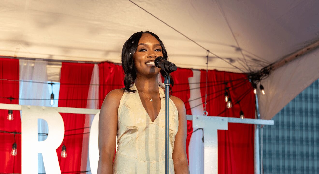 Woman speaking into microphone on stage with red curtains in background.
