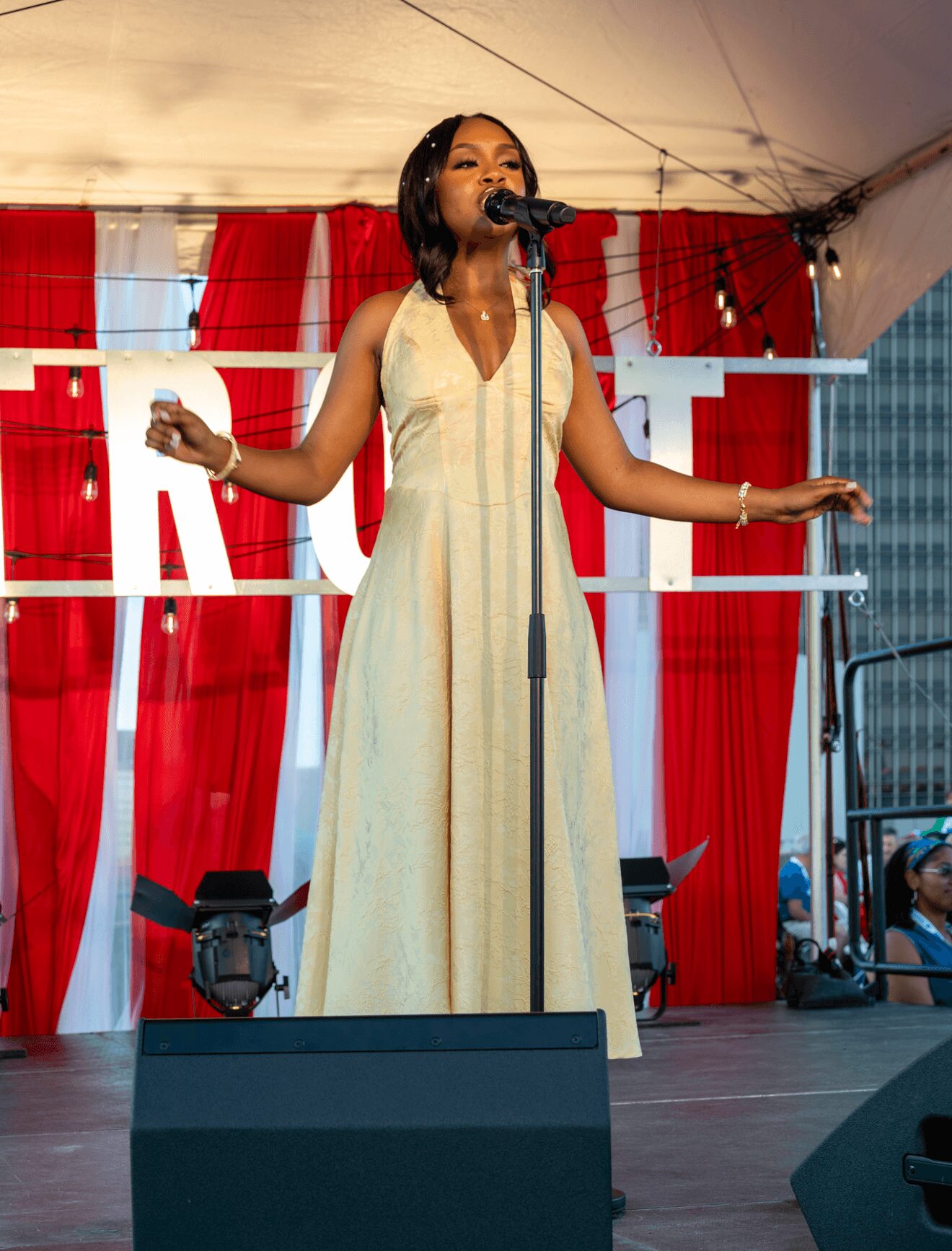 Woman singing on stage with red and white striped backdrop, wearing a long light-colored dress, arms outstretched.
