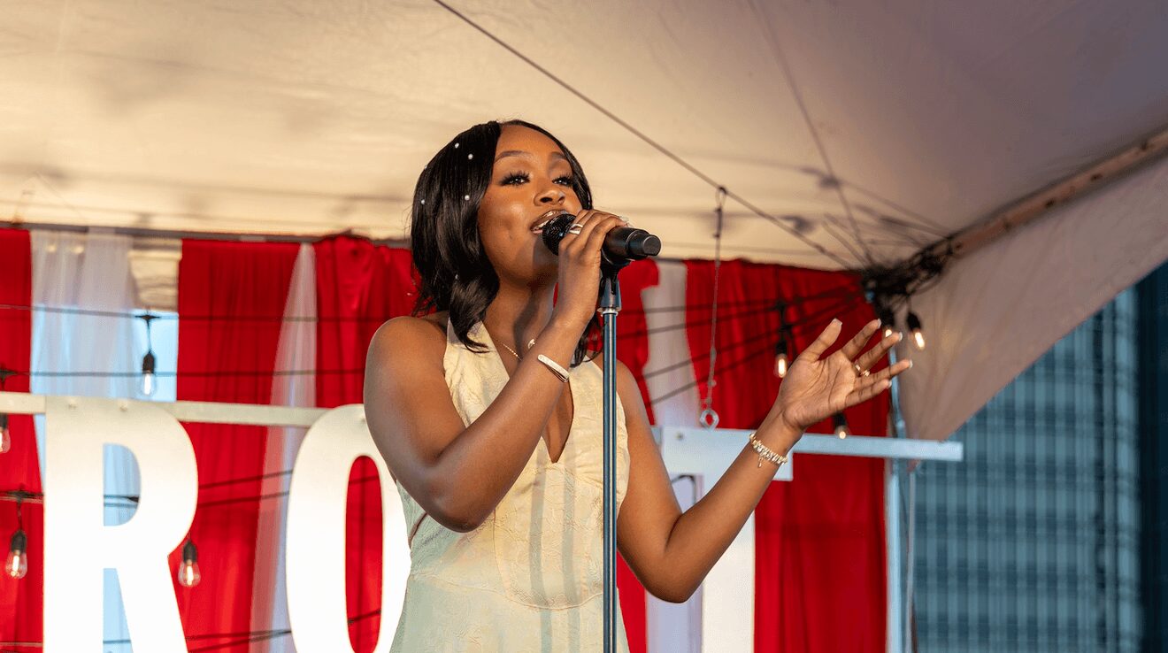 Woman singing into microphone on stage with red and white curtains behind her.