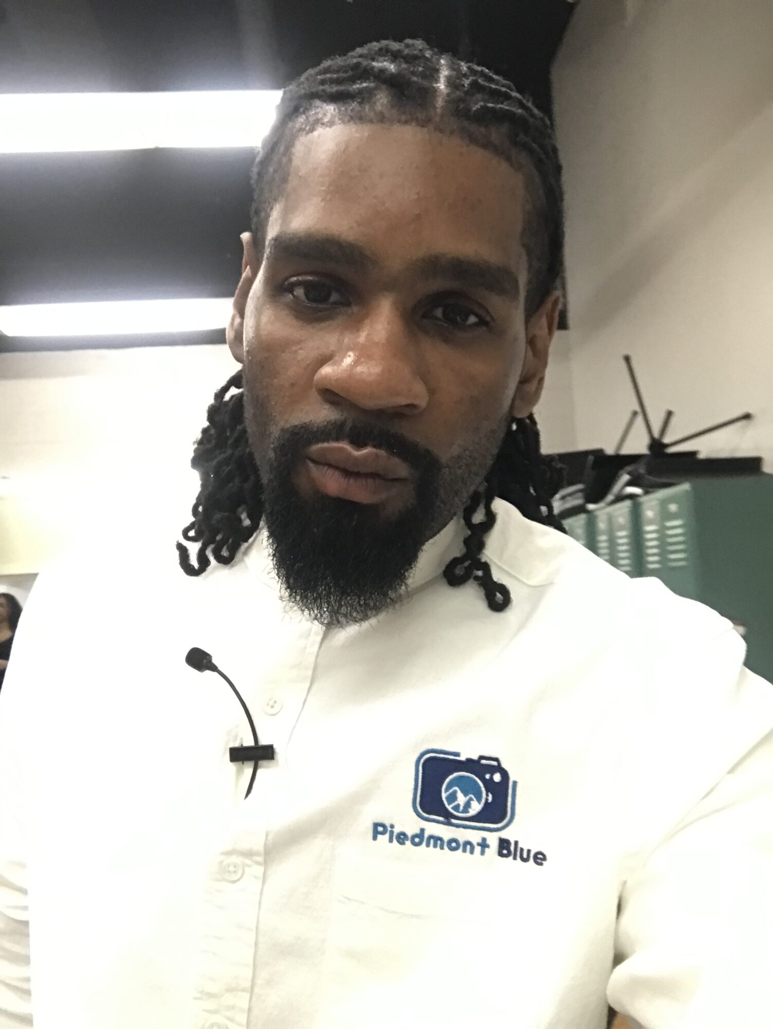 Man with braided hair and beard wearing a white shirt with a logo, indoors with fluorescent lights.