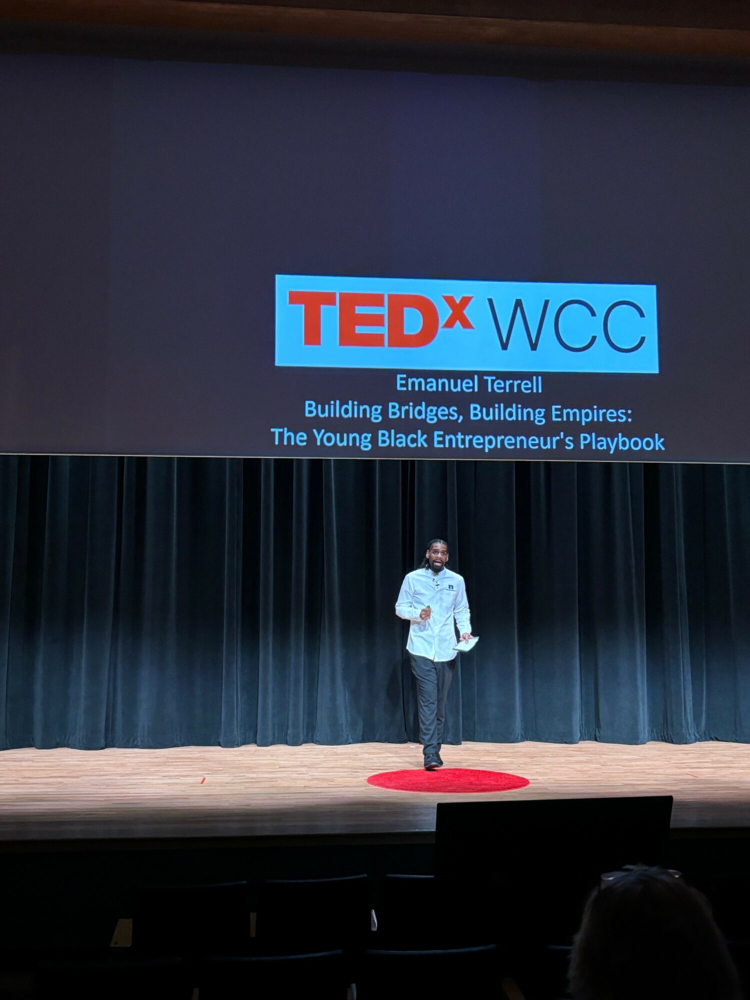 Person standing on stage with TEDx WCC logo and presentation title on screen behind, stage with black curtains, audience visible.