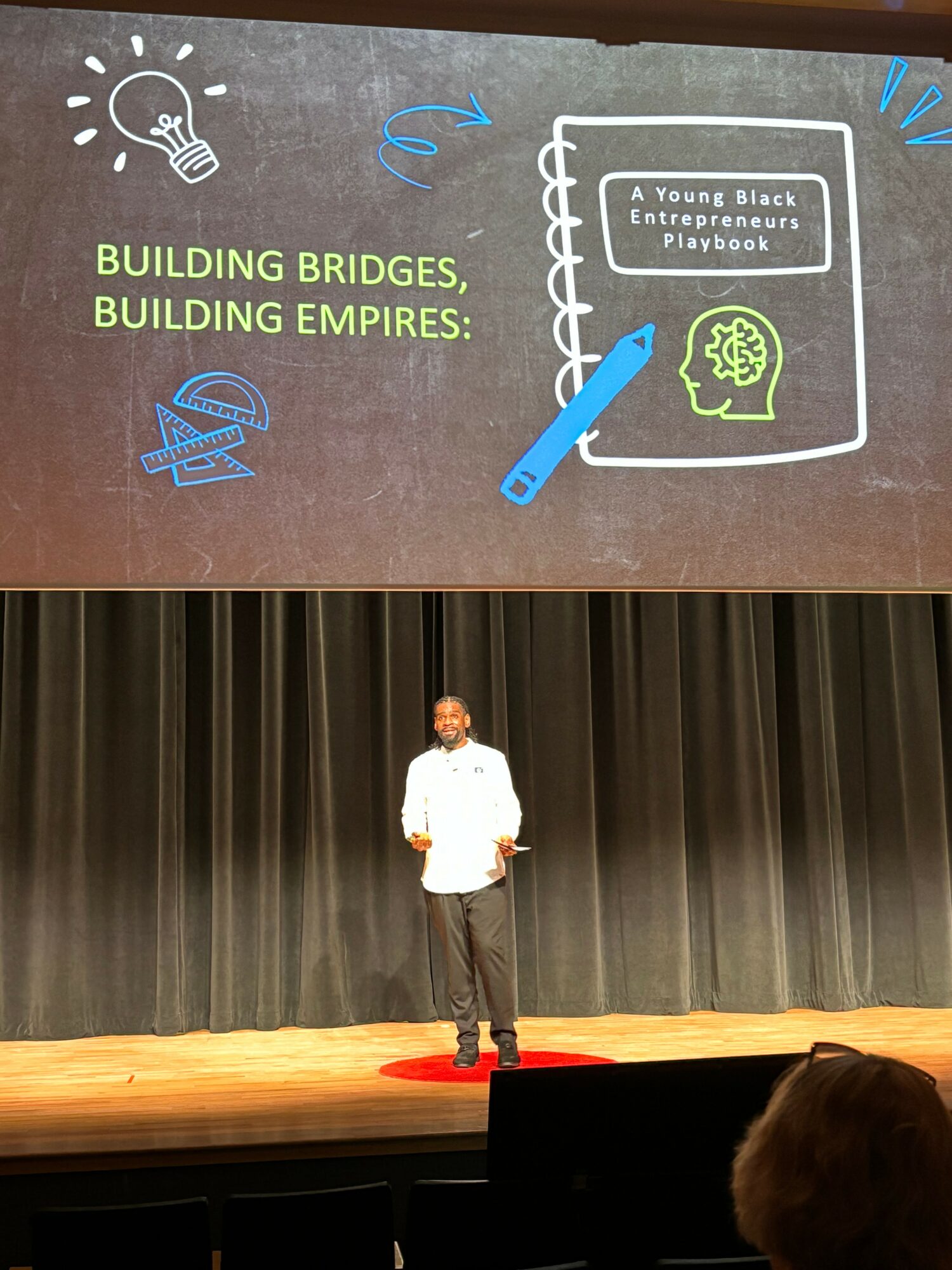 Person standing on stage with a large screen behind them displaying a book cover titled 'A Young Black Entrepreneurs Playbook' and the phrase 'Building Bridges, Building Empires'.