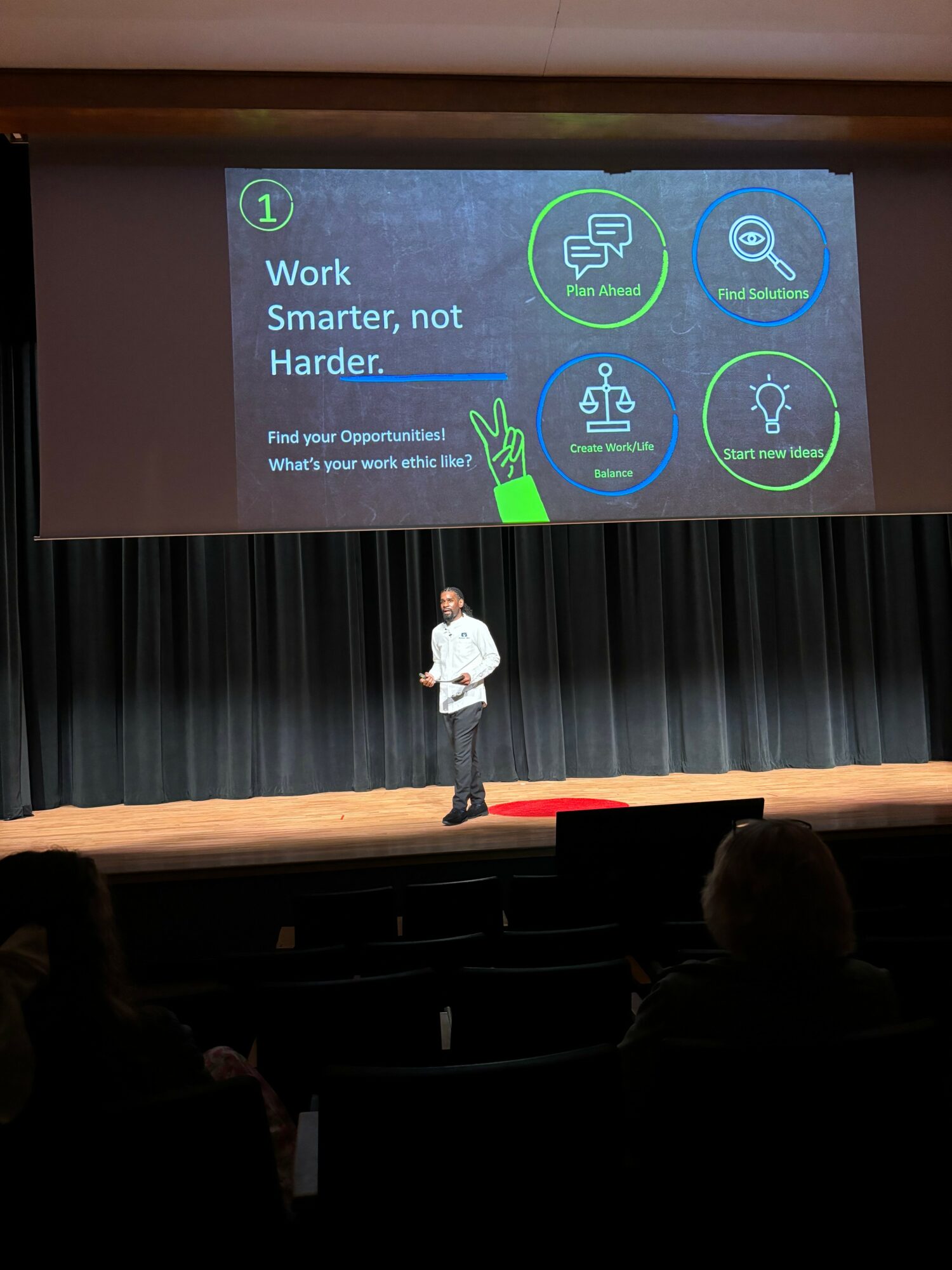 Person standing on stage in front of a large screen with icons and text, audience seated in front.