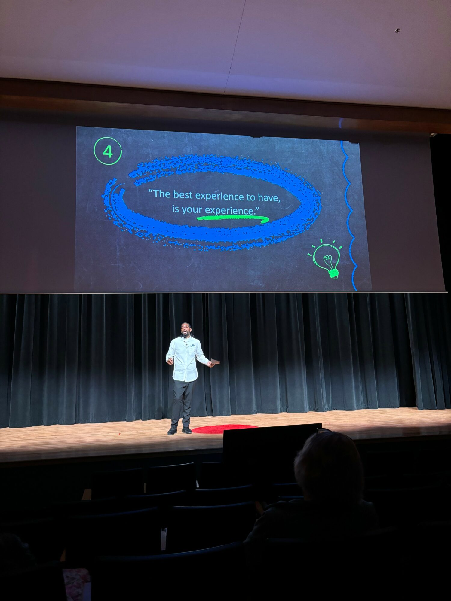 Person standing on stage in front of a large screen with a quote about experience, black curtain backdrop, auditorium seats visible.