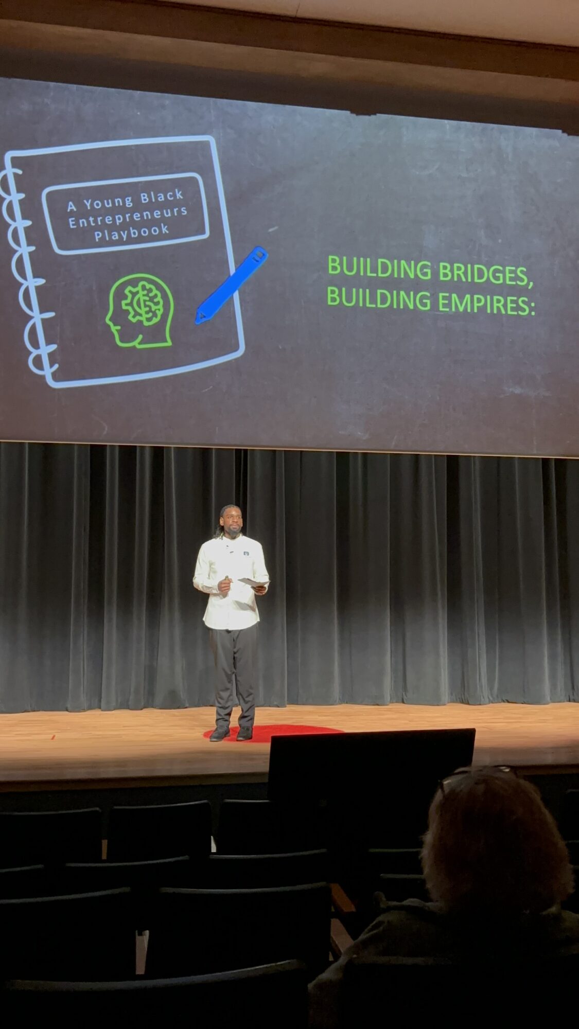 Person standing on stage with a presentation slide behind, showing a notebook with a brain icon and text about building bridges and empires.