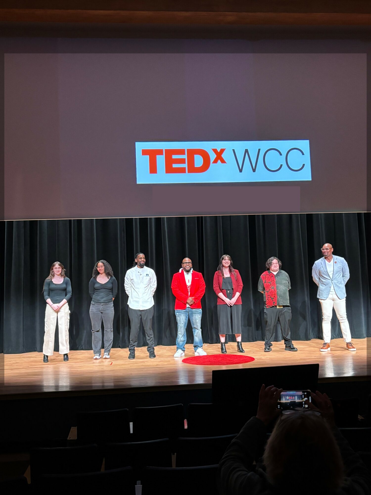 Seven people stand on stage in front of a TEDx WCC screen, with a person taking a photo in the foreground.