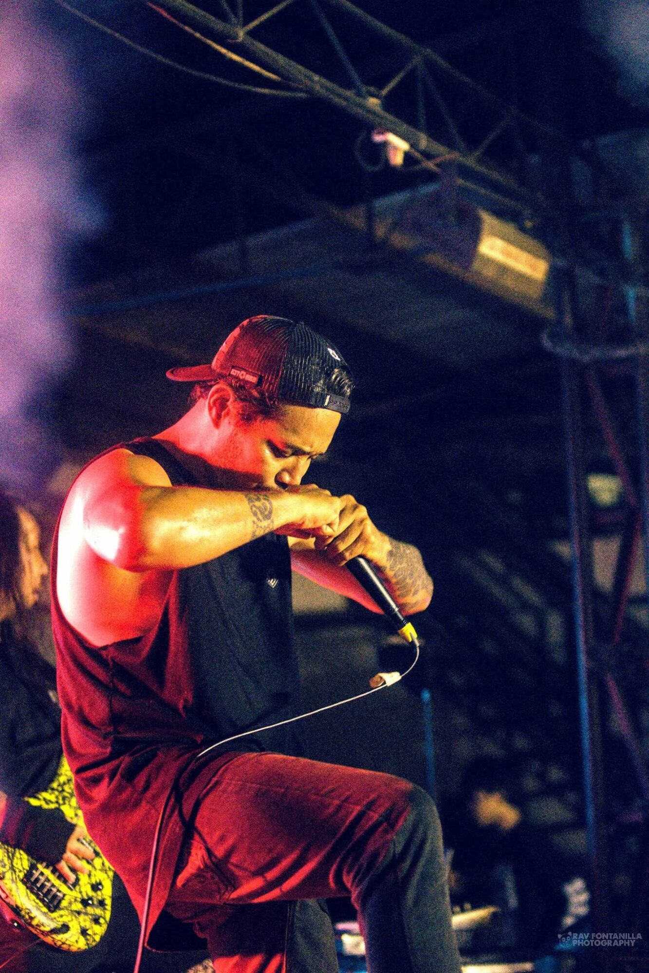 Person wearing a cap and sleeveless shirt holding a microphone on stage, with stage equipment overhead.