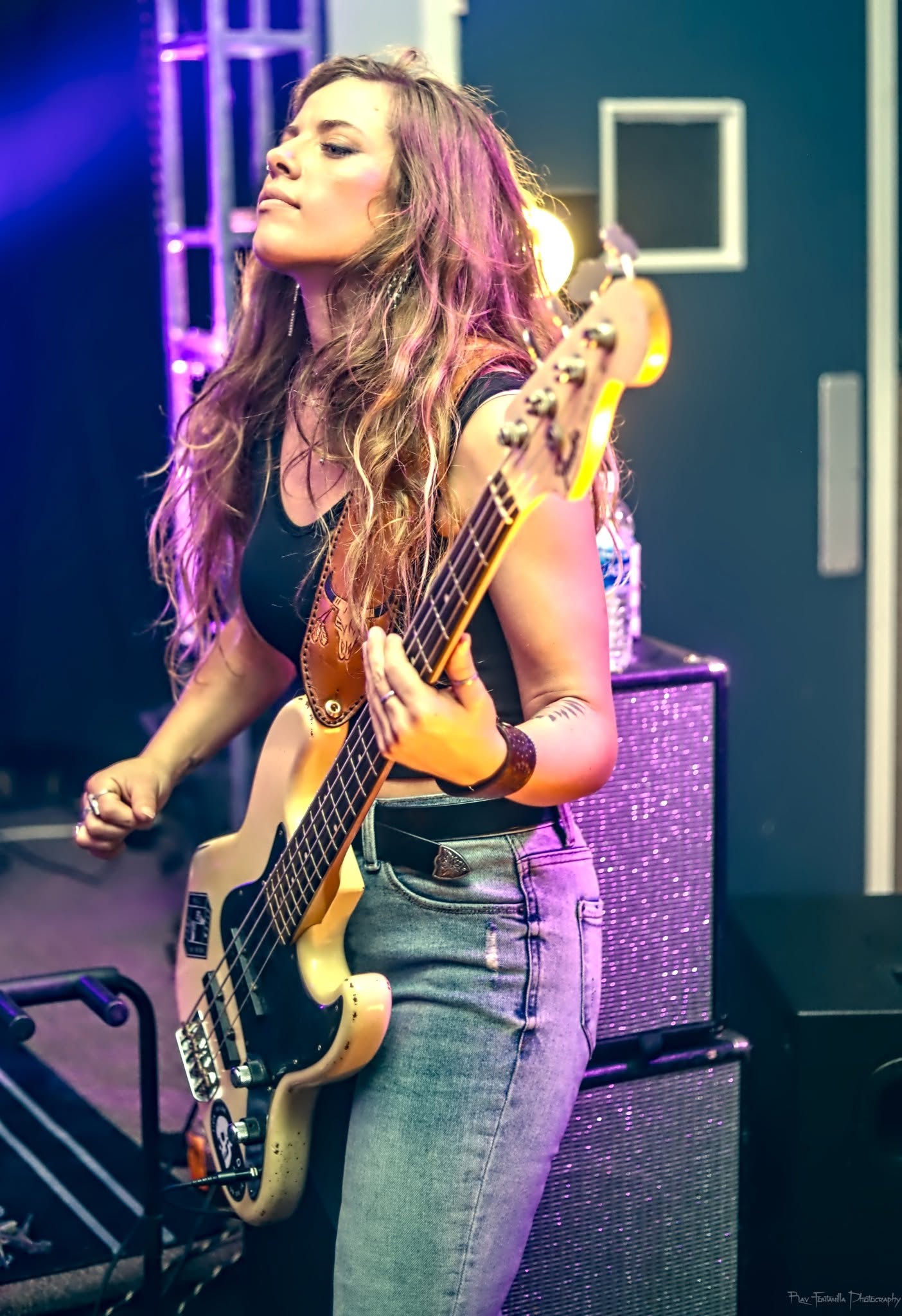 Young woman playing bass guitar on stage with purple lighting, wearing a black top and jeans.