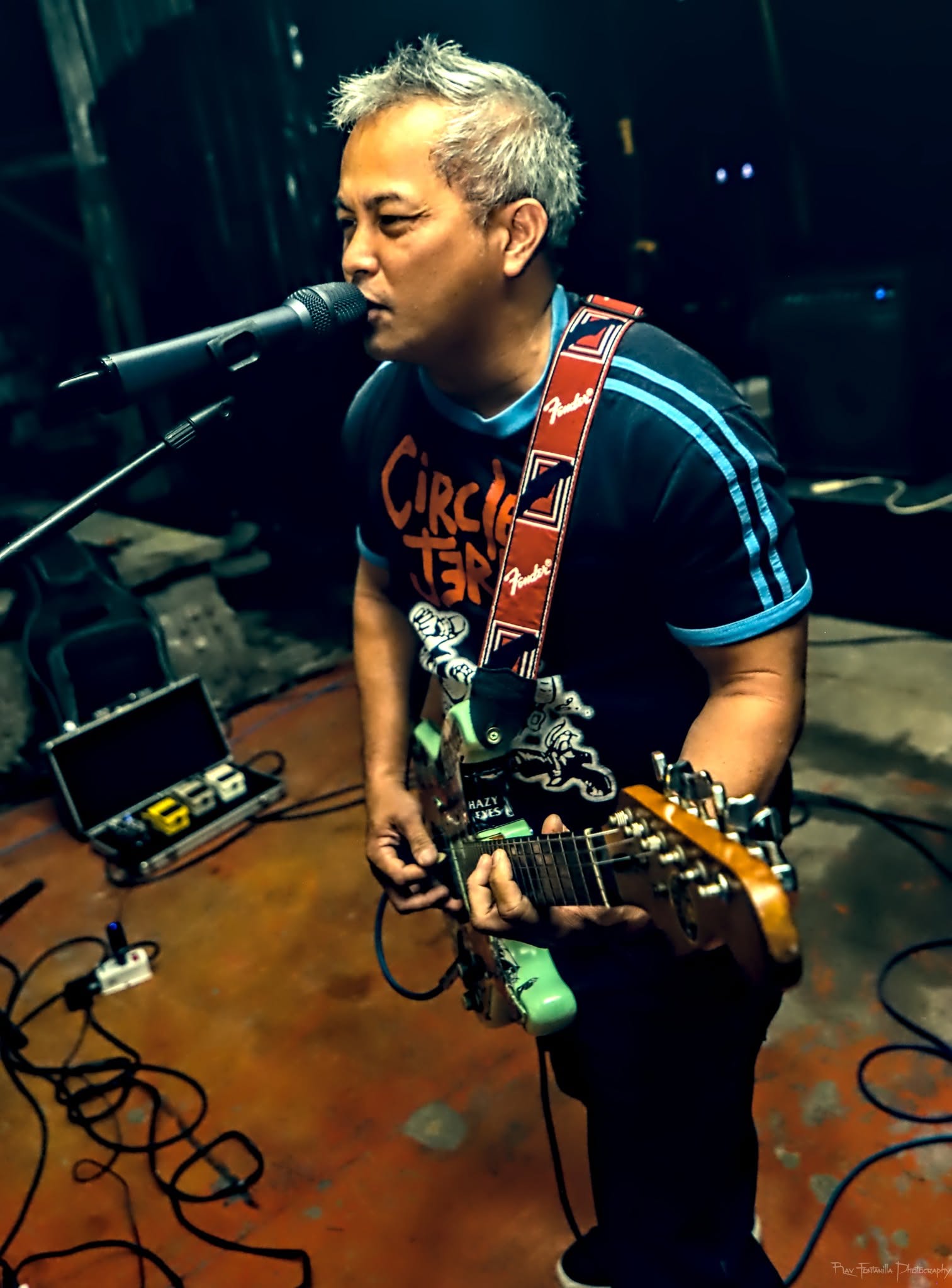 Person with short gray hair singing into a microphone while playing an electric guitar, wearing a striped shirt, in a dimly lit setting.