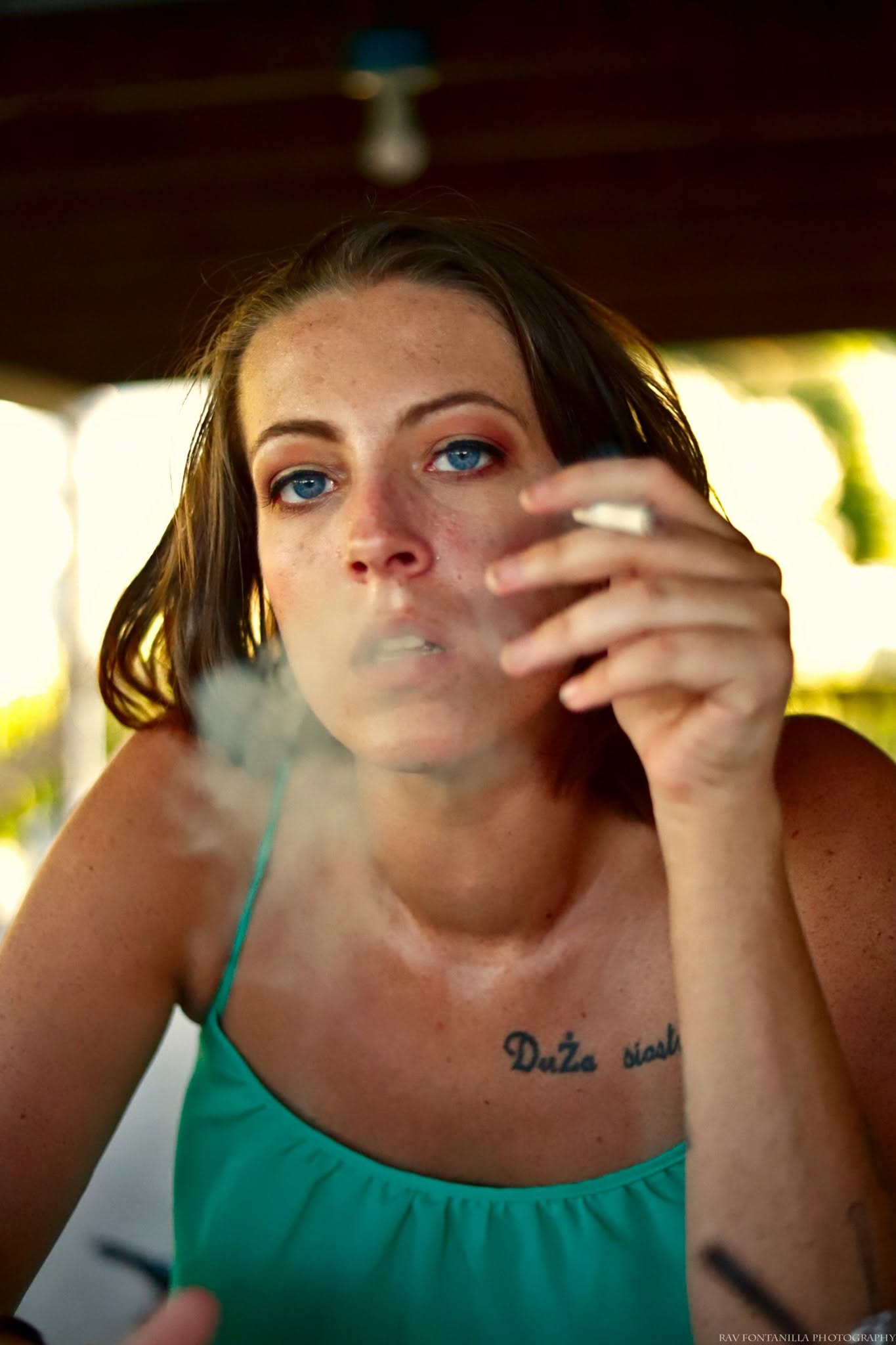 Woman with blue eyes and brown hair smokes, holding a cigarette near her face, with a tattoo on her chest, outdoors.