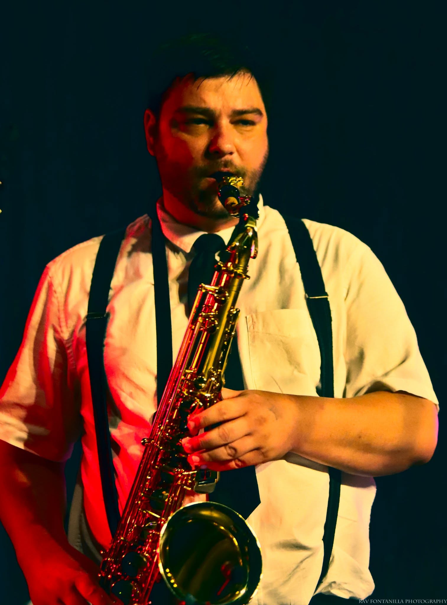 Person playing saxophone, wearing a white shirt with suspenders, against a dark background.